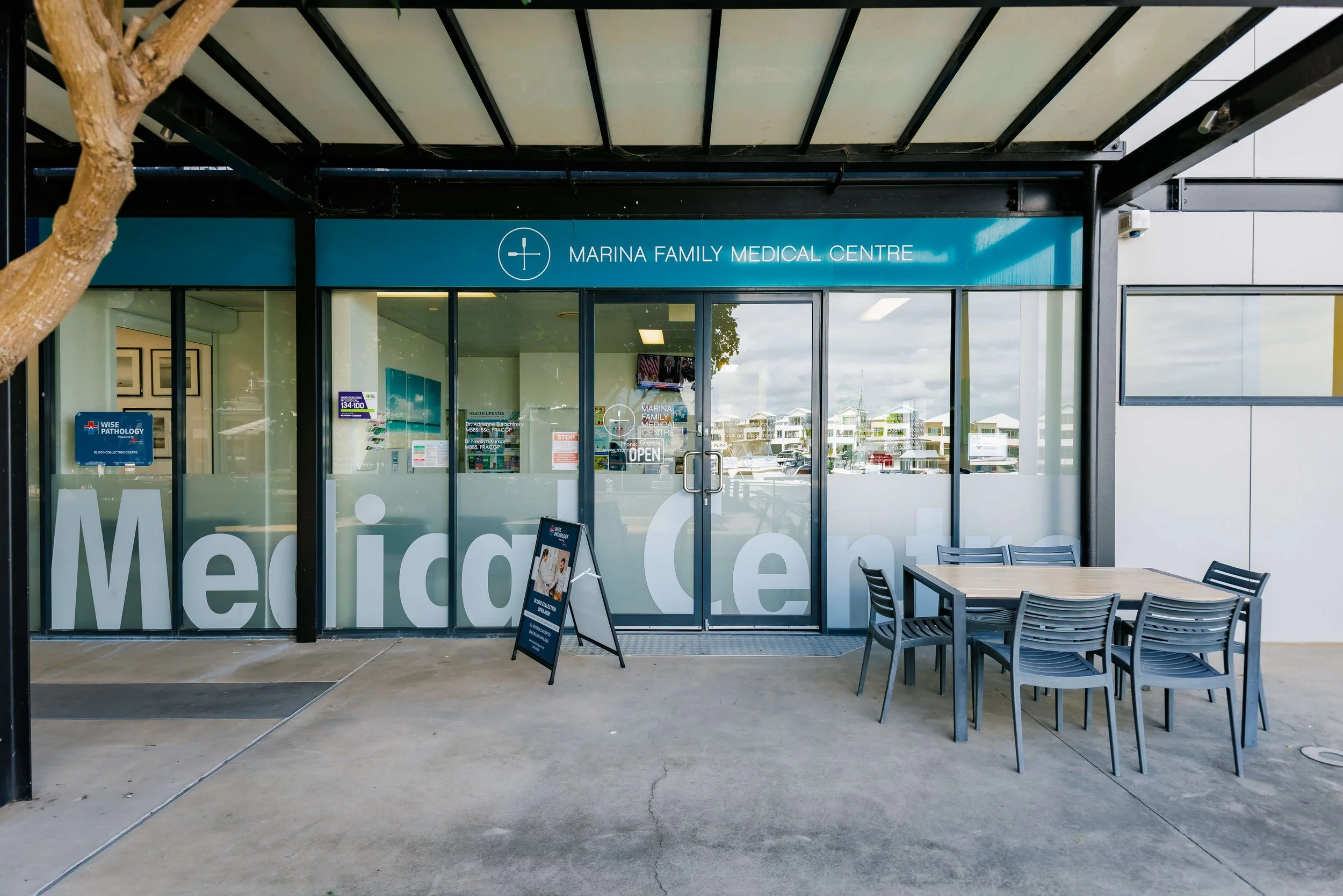Front entrance of Marina Family Medical Centre with glass doors, a blue sign, and outdoor seating area under a canopy.