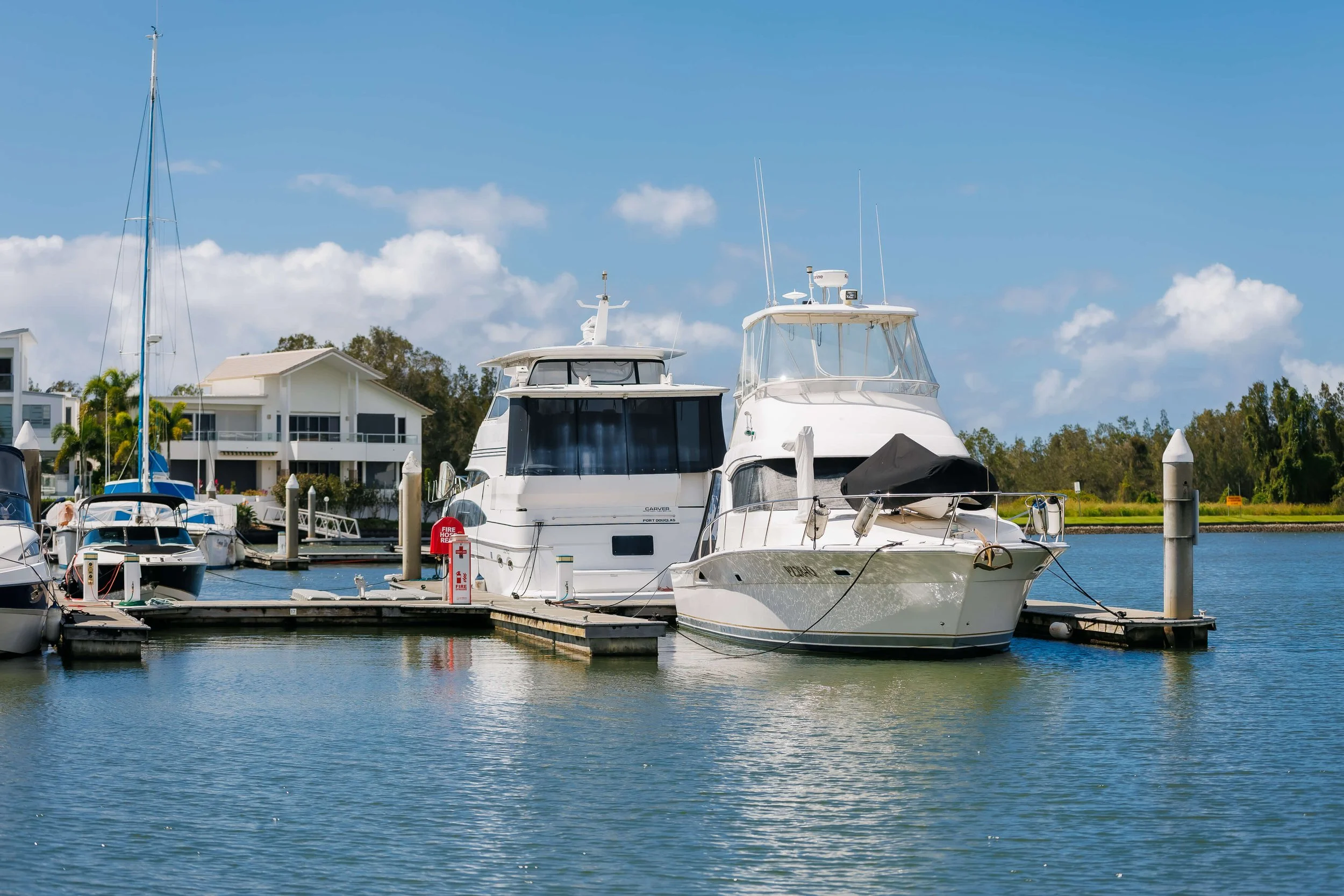 Boats docked at a marina on a sunny day, with white houses in the background and a blue sky.