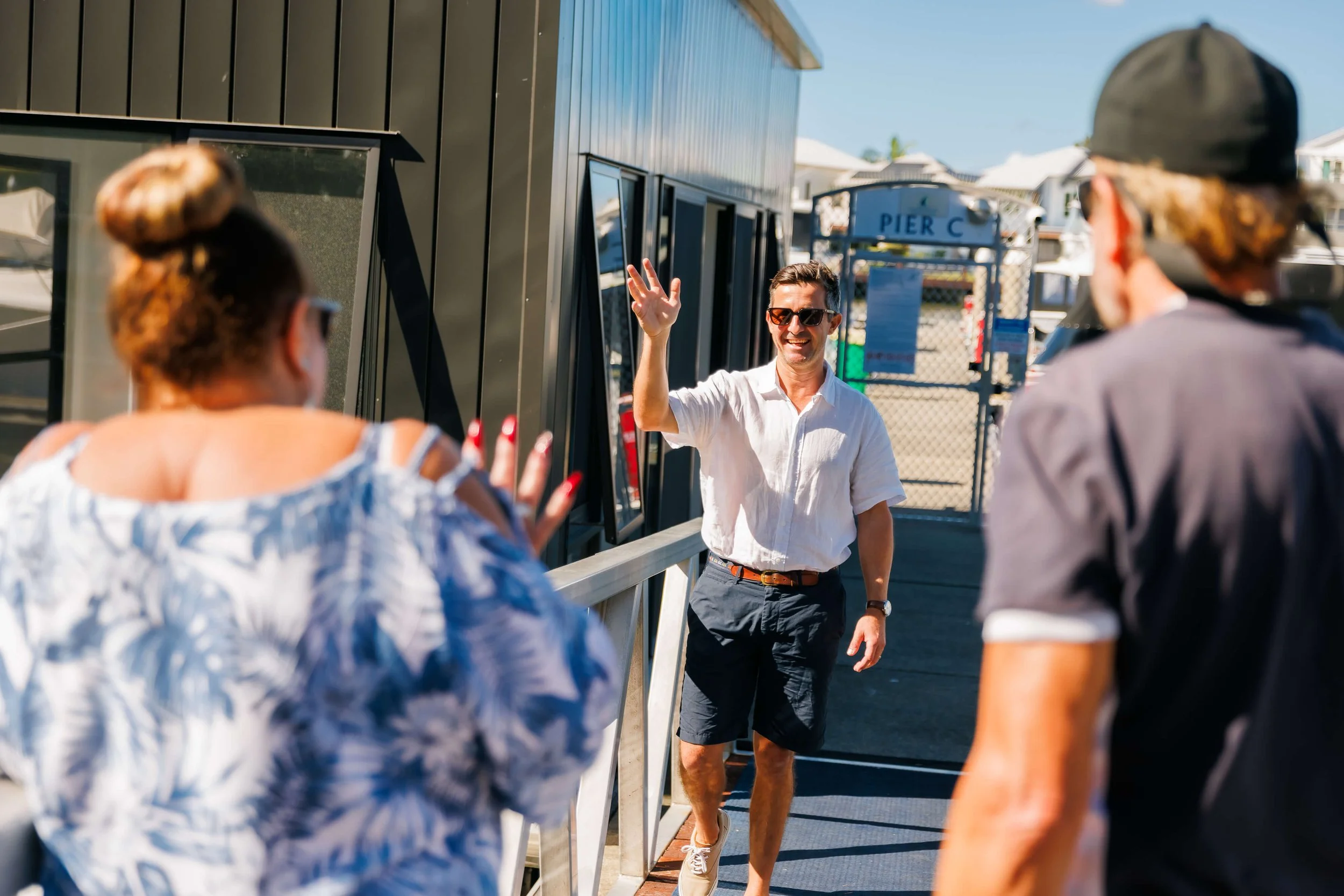 A man in sunglasses and a white shirt waves to a couple at a dock.