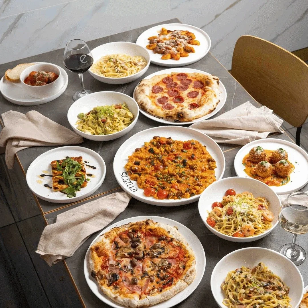 An assortment of Italian dishes on a table, including two pizzas, pasta dishes, meatballs, and a glass of red wine.