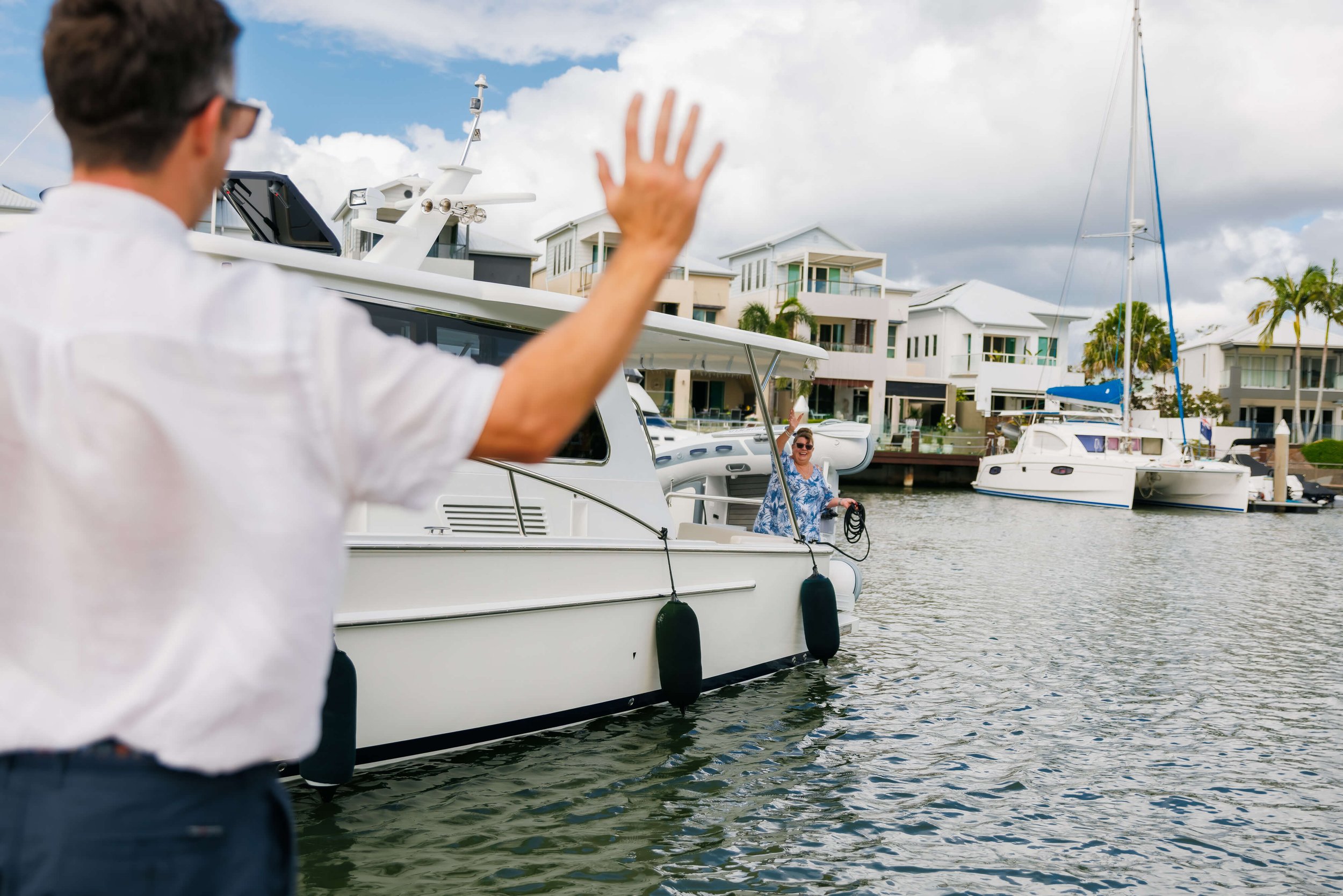 A man in a white shirt waving at a woman on a docked boat at a marina, with modern residential buildings and palm trees in the background.