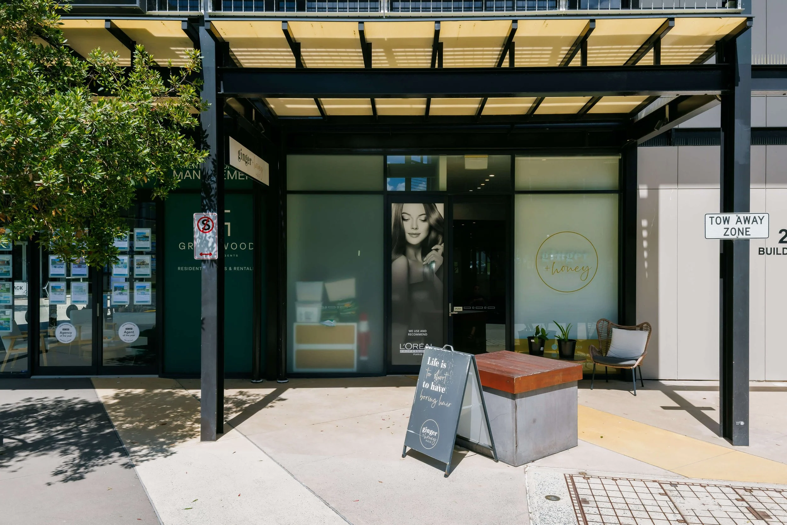 Exterior view of a storefront with glass doors and windows, outdoor seating with chairs and tables, and greenery including trees and potted plants.