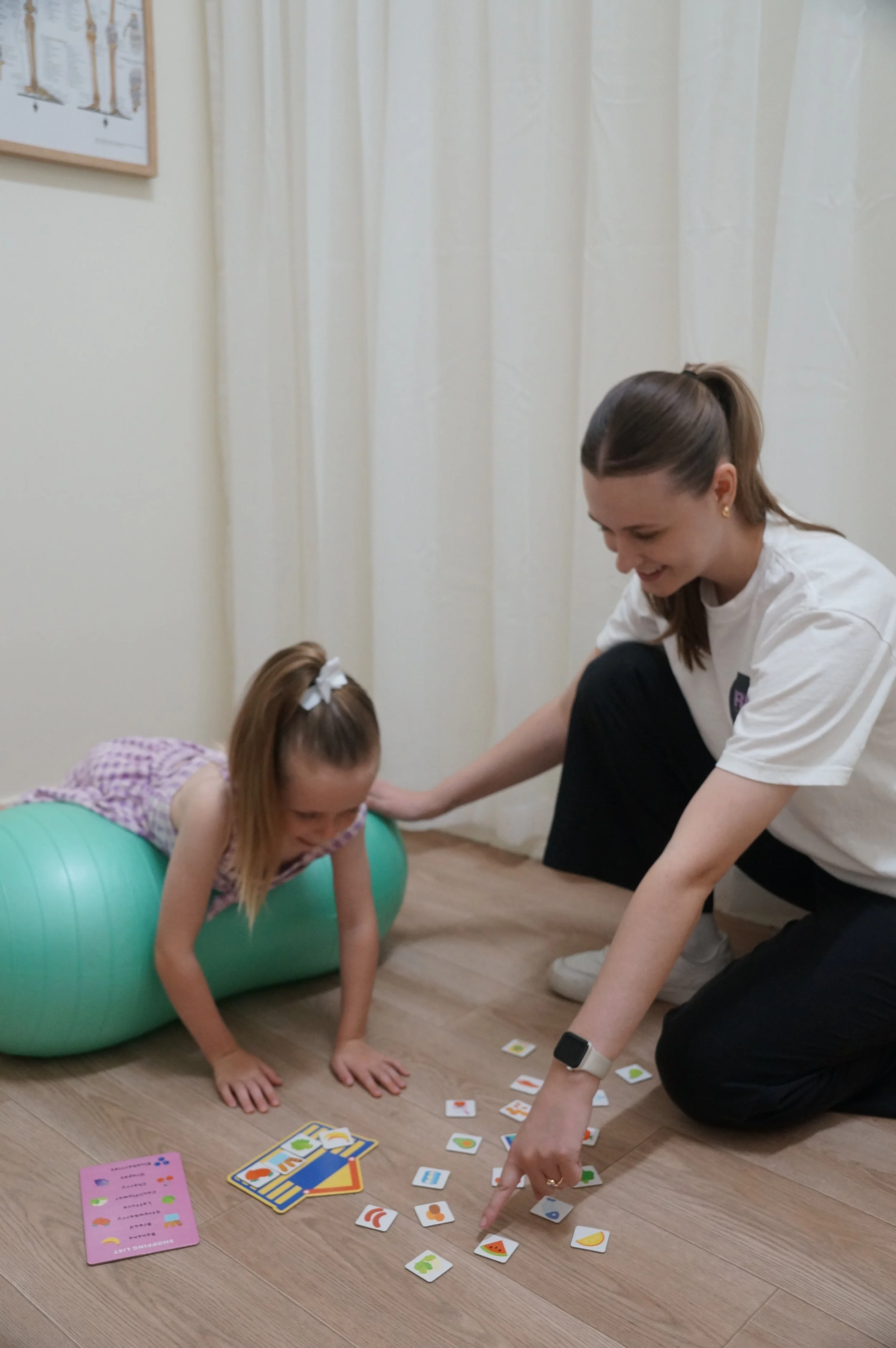 A Paediatric Physiotherapist (Makenzie Burridge) and a young girl playing a card game on the floor in a room with white curtains. The girl is on an exercise ball, and the woman is kneeling, both engaged with game cards.
