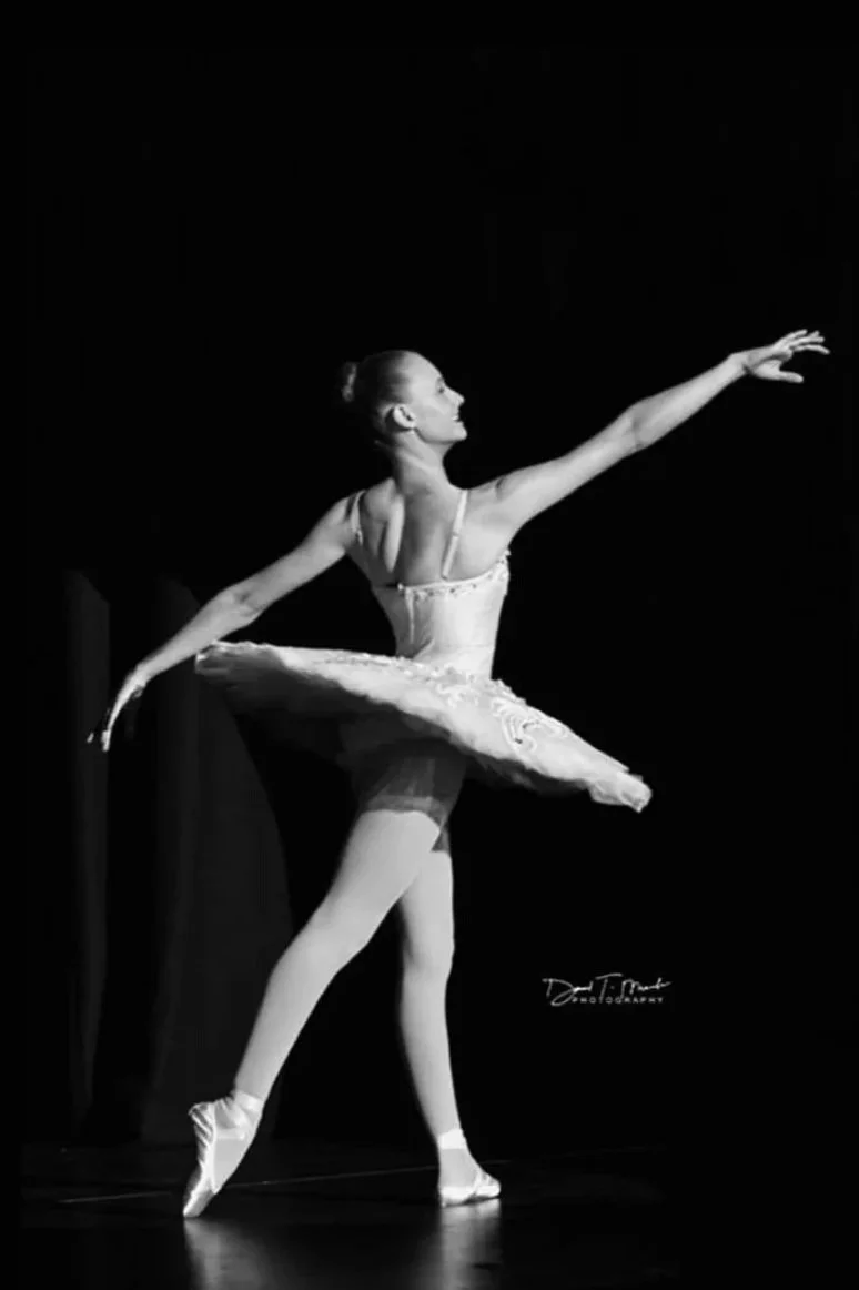 Black and white photo of a female ballet dancer (Makenzie Burridge) in a tutu, standing on stage with one arm extended and the other arm out to the side.
