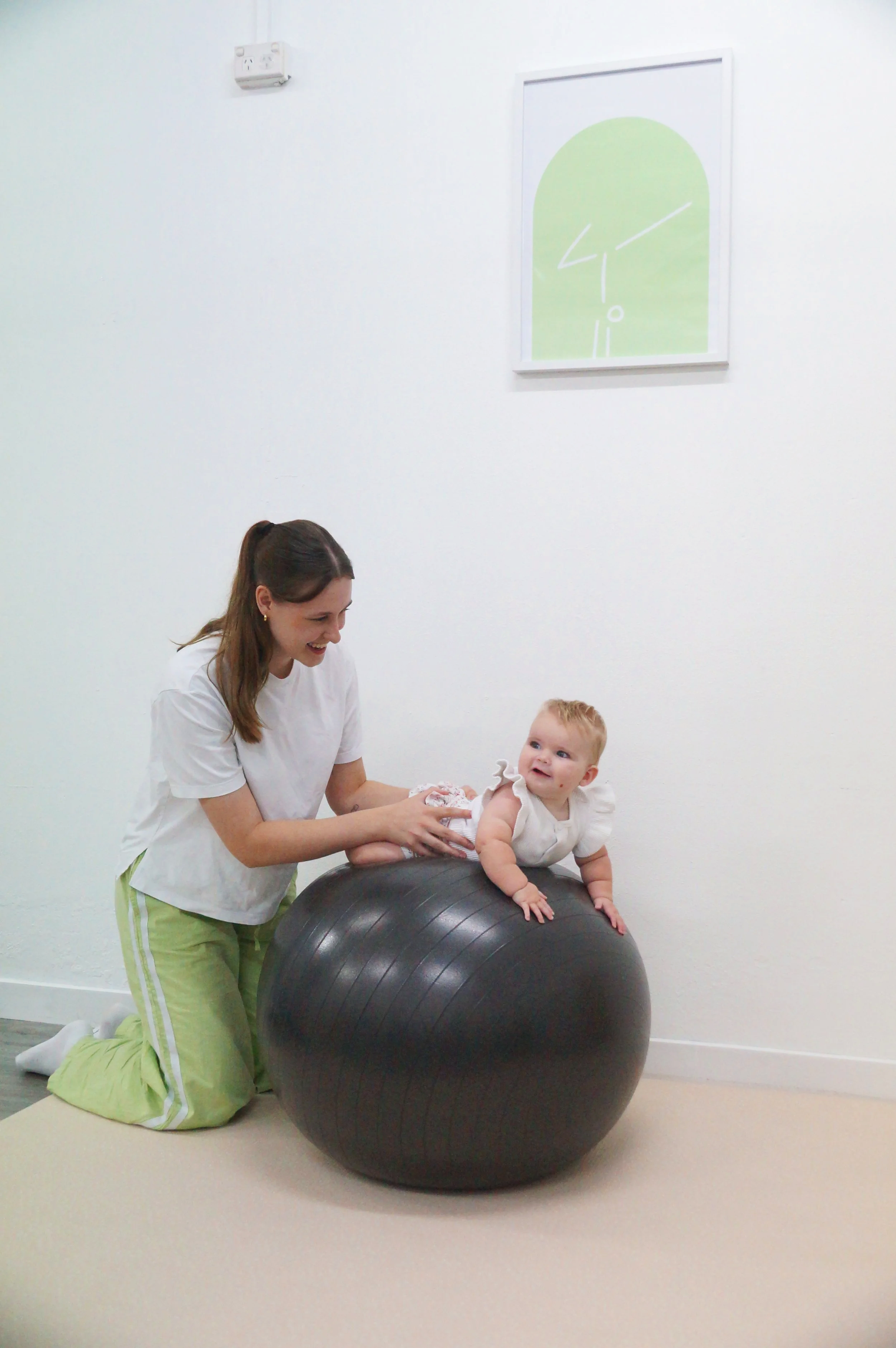 A Physiotherapist (Makenzie Burridge) is kneeling on the floor holding a baby on a large black exercise ball in a white room with a minimalistic painting on the wall.