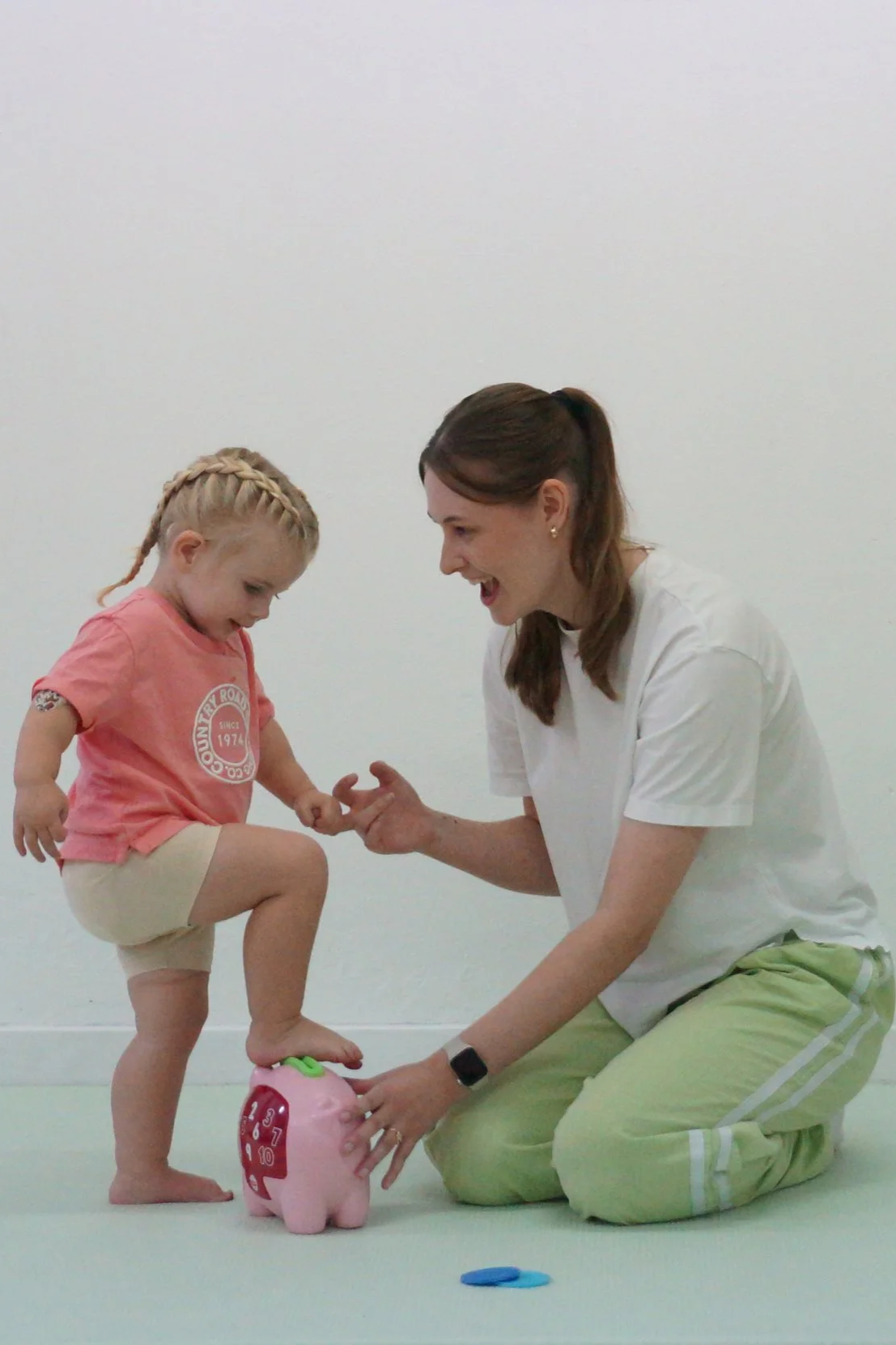 A young girl stepping on a piggy bank toy, with a Physiotherapist (Makenzie Burridge) in a white t-shirt and green pants kneeling nearby, both engaging in a playful activity challenging balance.