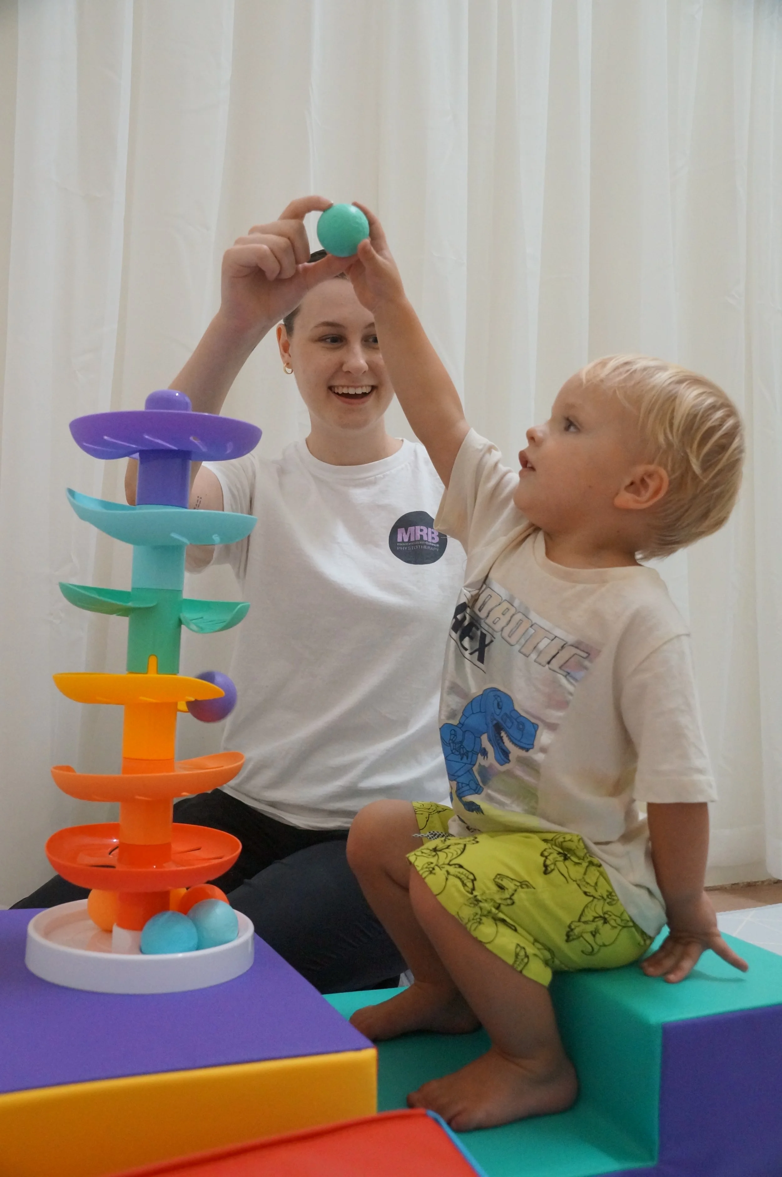 A Physiotherapist (Makenzie Burridge) and a young boy playing a marble game with a colourful marble tower in a room with white curtains.