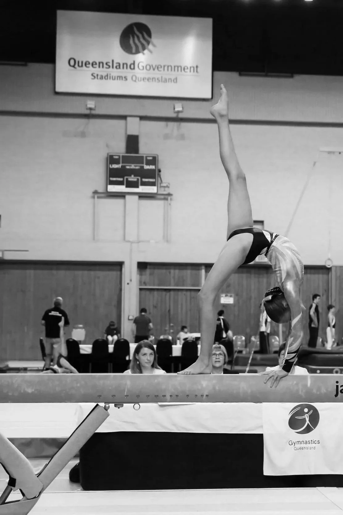 A female gymnast (Makenzie Burridge) performing a handstand on a balance beam during a gymnastics event at a Queensland Gymnastics competition, with spectators and officials in the background.