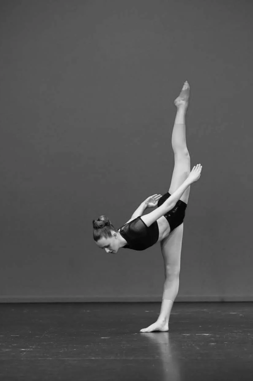 A female dancer (Makenzie Burridge) performing a balancing pose on one foot with her other leg extended vertically upward, on a stage against a plain background.