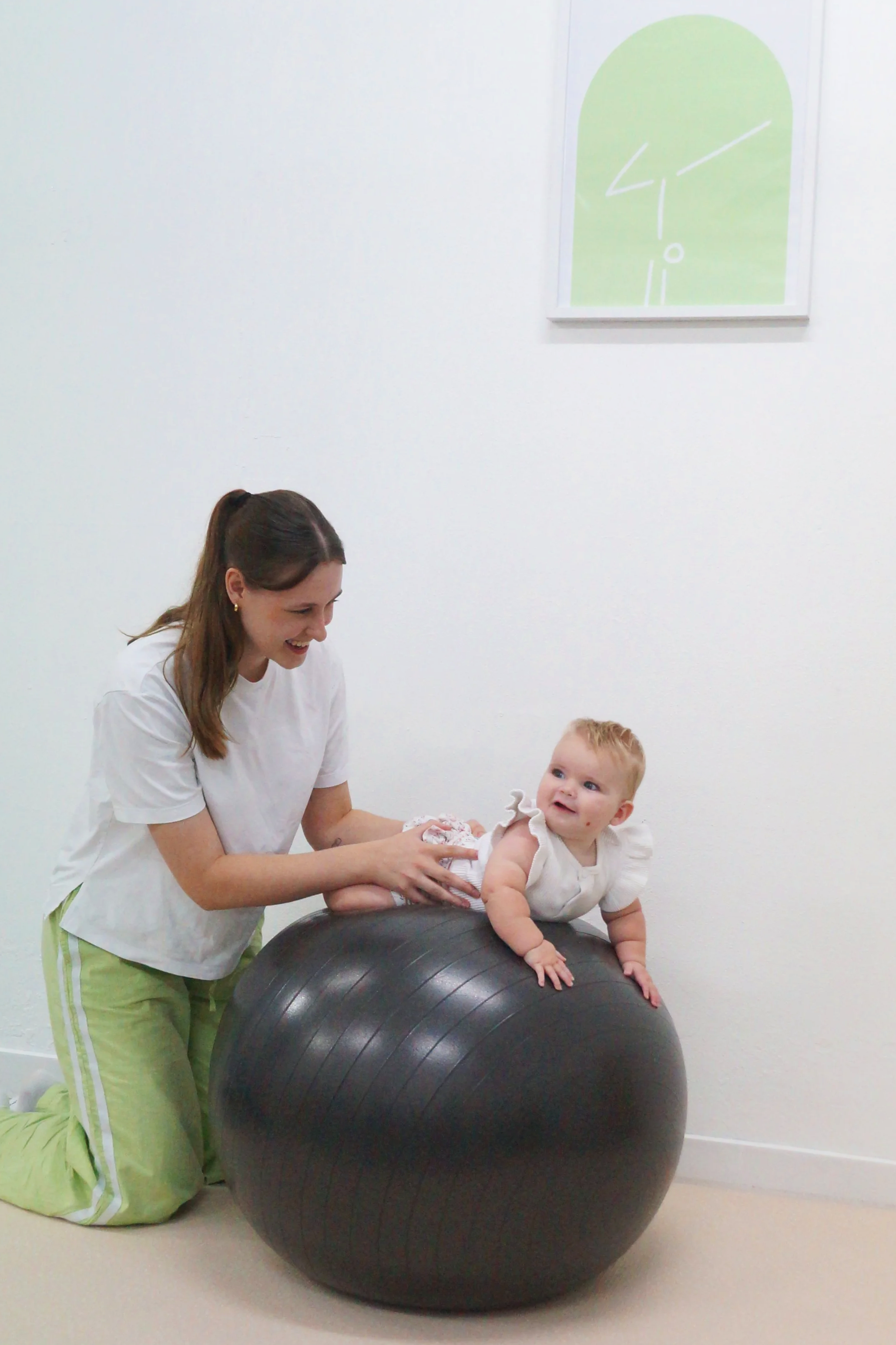 A Physiotherapist (Makenzie Burridge), wearing a white t-shirt and light green pants, is smiling while helping a baby to crawl on a black exercise ball in a white room with minimalist decor. There is a green abstract art piece on the wall.