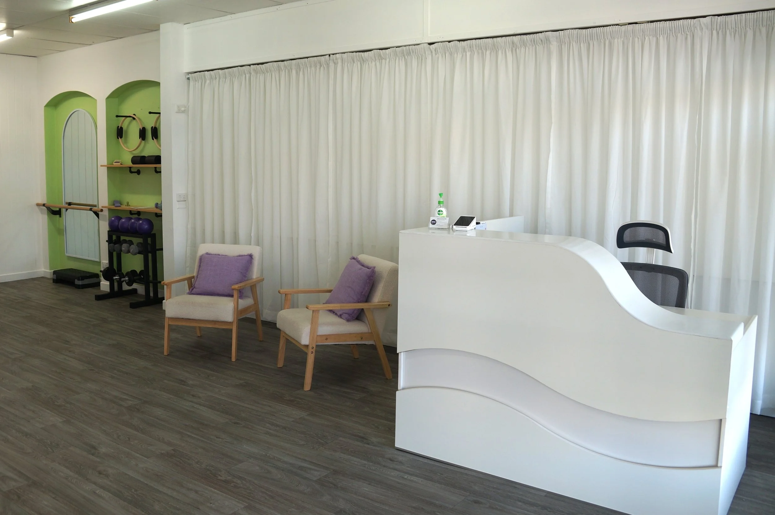 Interior view of a modern waiting area with chairs, a white reception desk, and a curtain. There is gym equipment on the left side and a wooden floor.