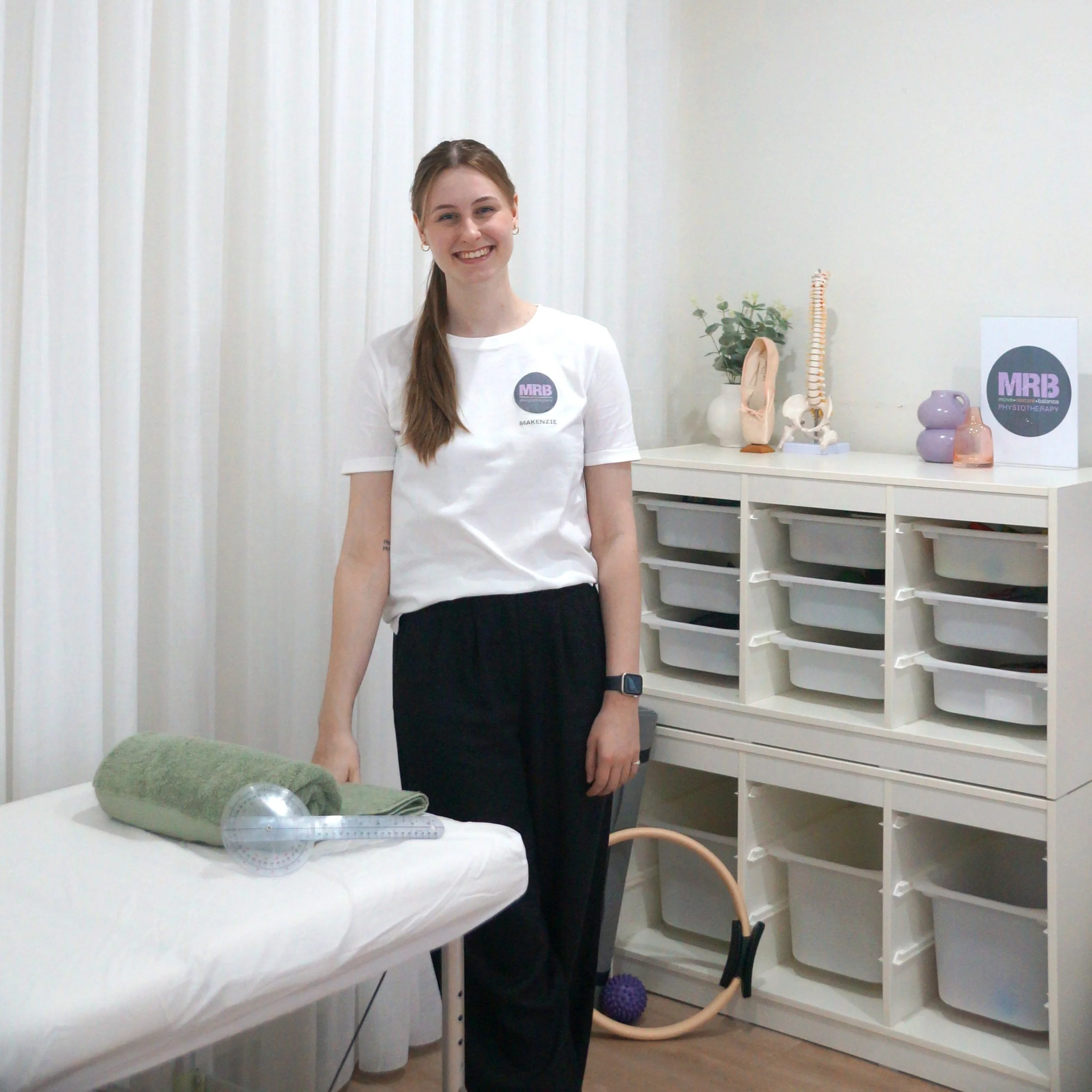 Smiling woman who is a Physiotherapist (Makenzie Burridge) in a white t-shirt with a logo and the name 'MAKENZIE' standing in a medical or therapy room, with medical models and equipment on the table and shelves behind her.