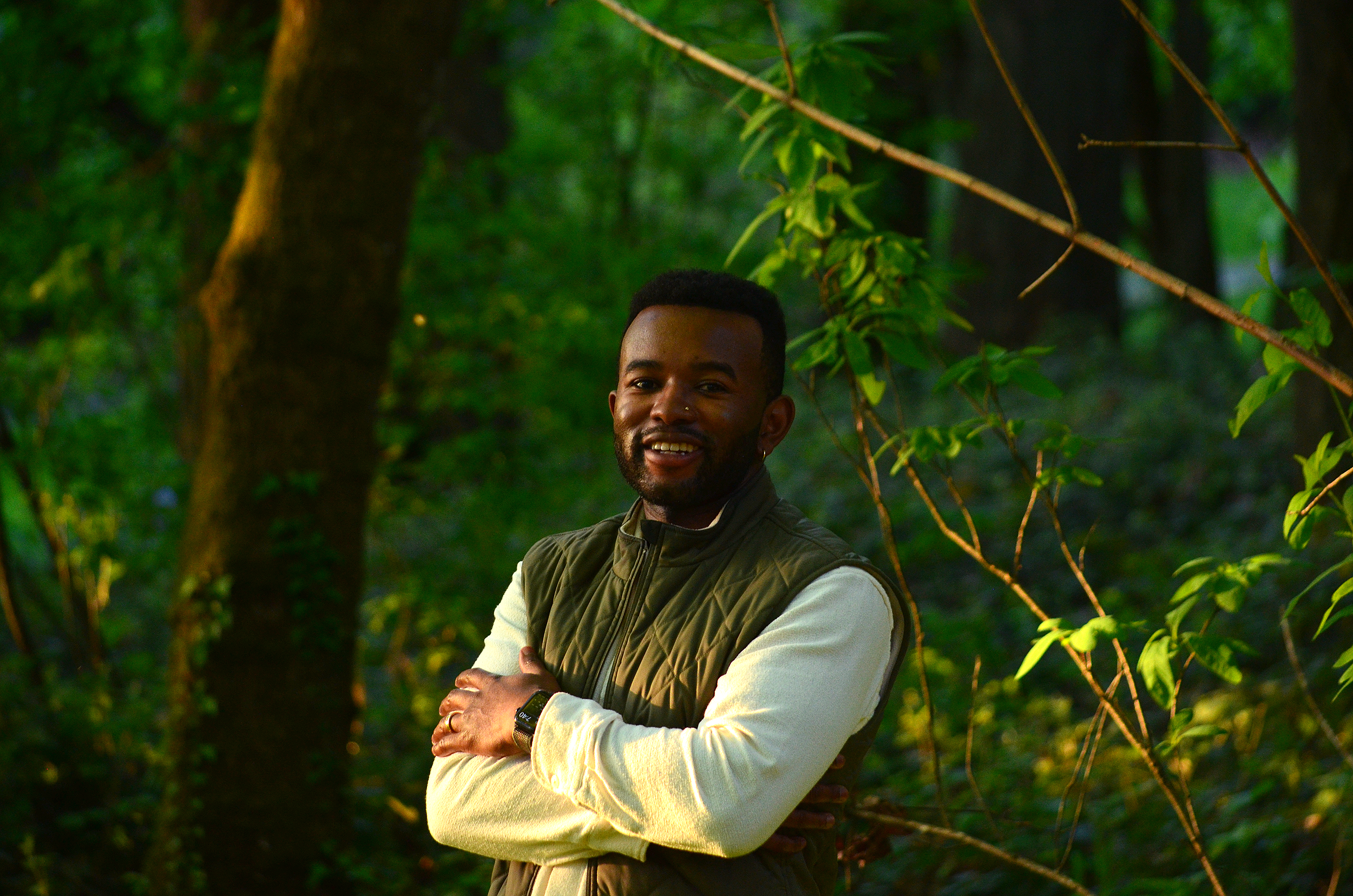 A man with dark skin, short black hair, and a beard stands outdoors among green trees, smiling with arms crossed, wearing a cream long-sleeve shirt and a green quilted vest.