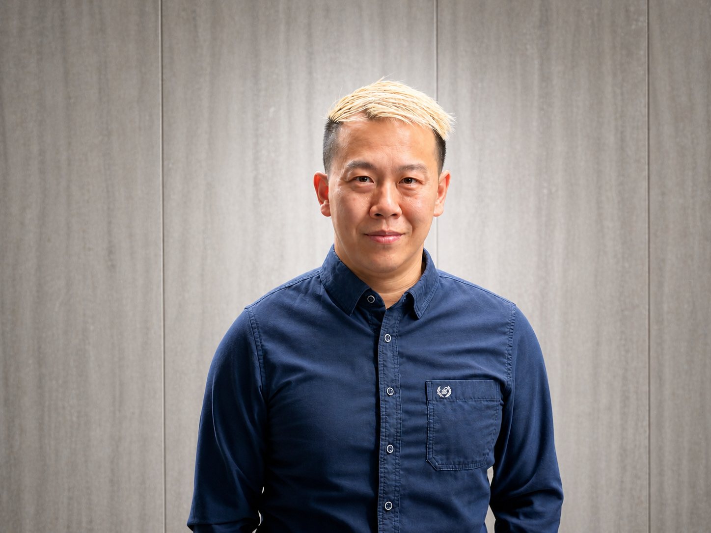 A man with short blonde hair and light skin wearing a dark blue button-up shirt, standing in front of a plain, light-colored wall.