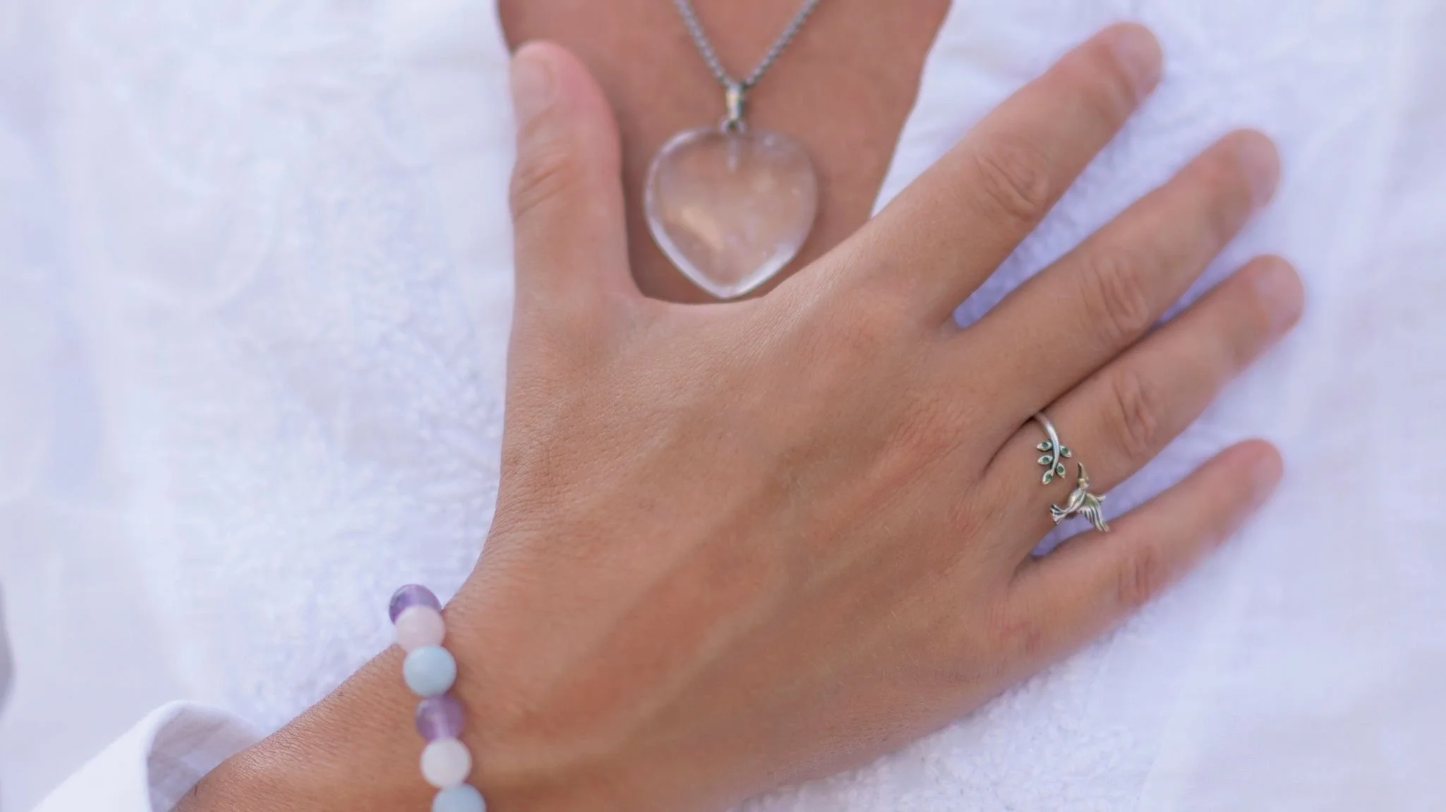 Mano con collar de cristal en forma de corazón y anillo de plata, sobre superficie blanca.