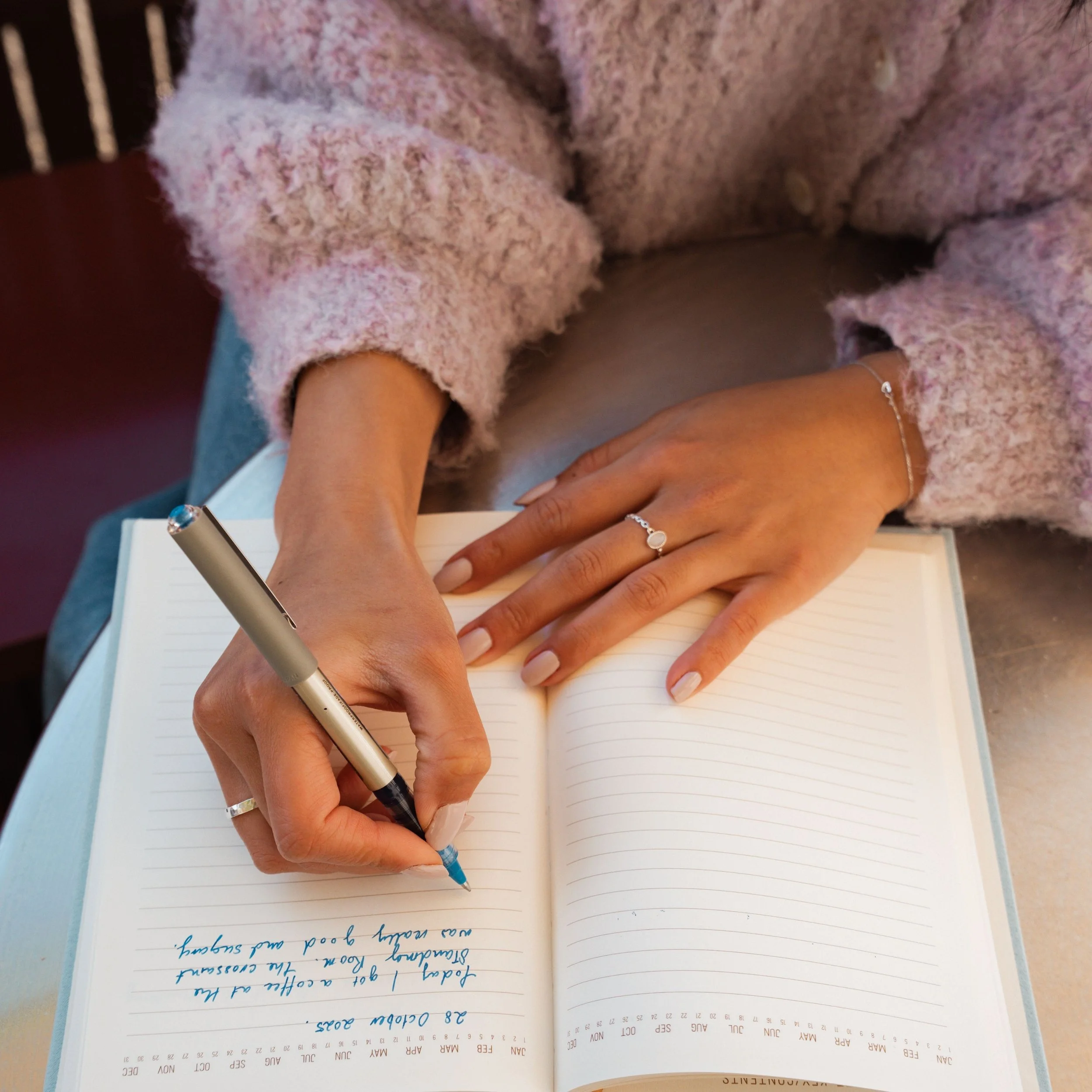 Una mano escribe en un cuaderno, con un bolígrafo plateado, en una hoja con escritura en color azul. La mano lleva un anillo y una pulsera plateada, y hay otro anillo en el dedo medio. La página del cuaderno tiene líneas horizontales y un calendario en la esquina inferior derecha. La imagen está ligeramente borrosa y muestra parte del brazo y la mano.
