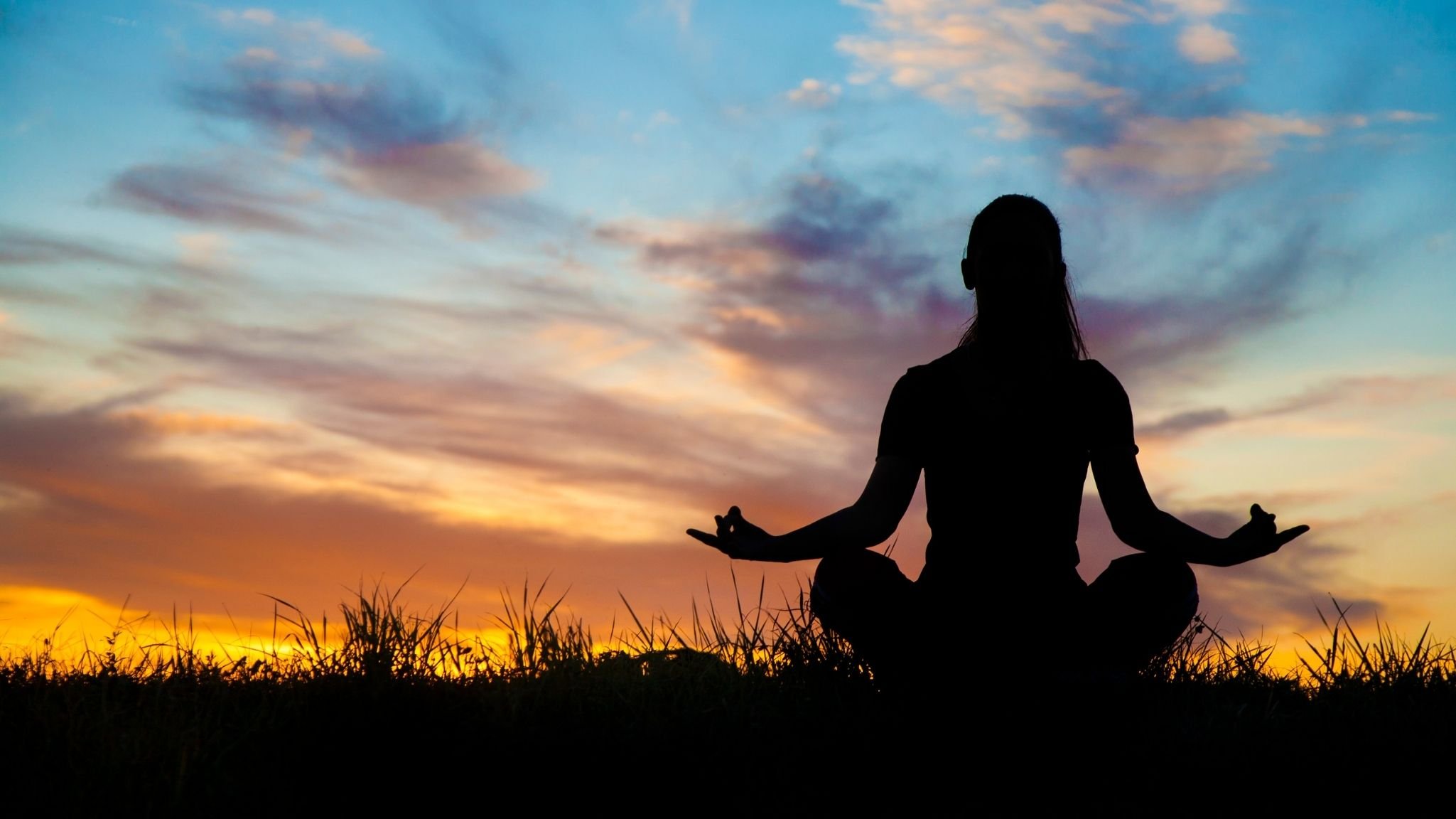Persona practicando yoga o meditación al atardecer, sentada en postura de loto con las manos en mudra en un campo