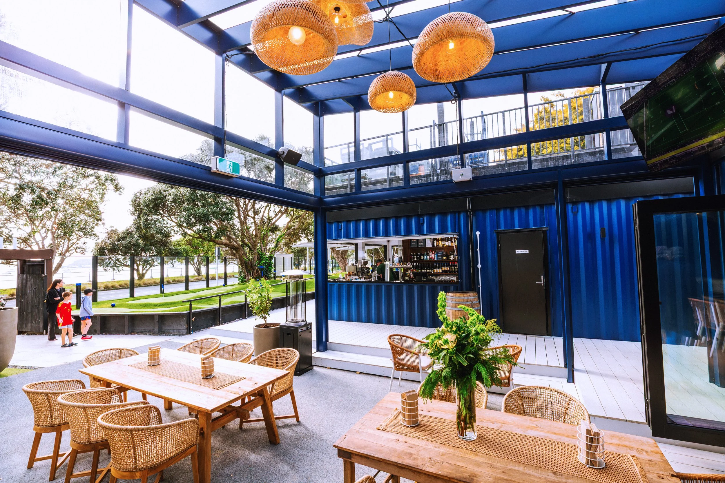 Interior of a modern restaurant with a glass ceiling and blue metal framing, surrounded by large windows showing trees outside. Woven pendant lights hang from the ceiling, and wicker chairs and wooden tables are arranged inside. There is an outdoor seating area with greenery and children playing in the background.
