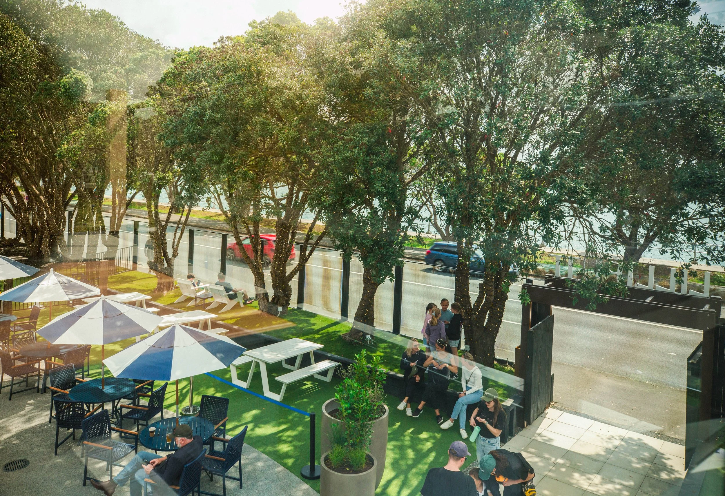 Outdoor patio with tables, umbrellas, and people sitting and standing by large trees decorated with string lights, near a street with cars passing by.