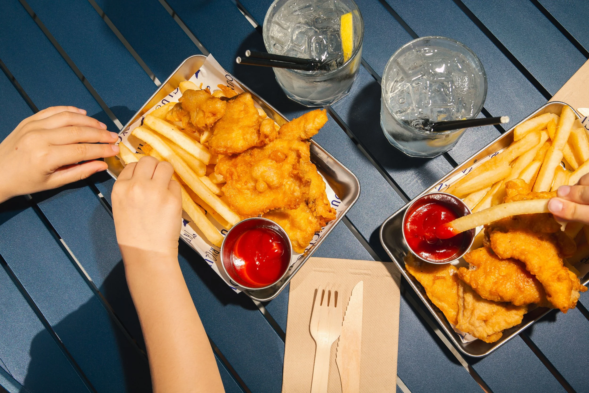 Two hands reaching for a tray of fried chicken and French fries on a blue table, with two glasses of ice water with lemon and straws.