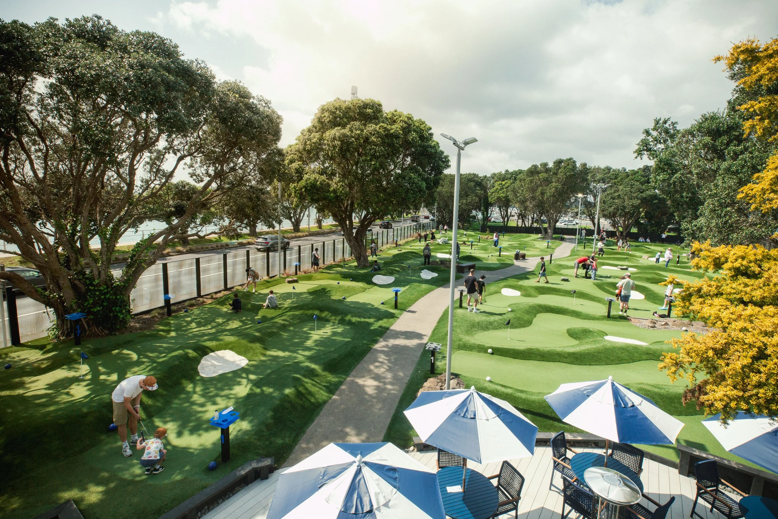A mini golf course with multiple holes, surrounded by green grass and large trees. People are playing, and there are umbrellas shading seating areas, with overcast skies overhead.