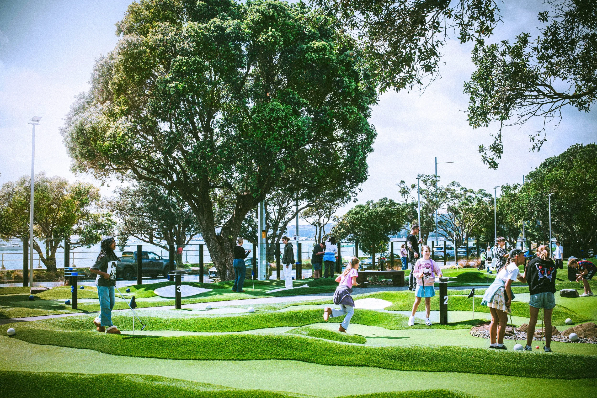 People playing mini golf in a park with large green trees and a waterfront in the background.