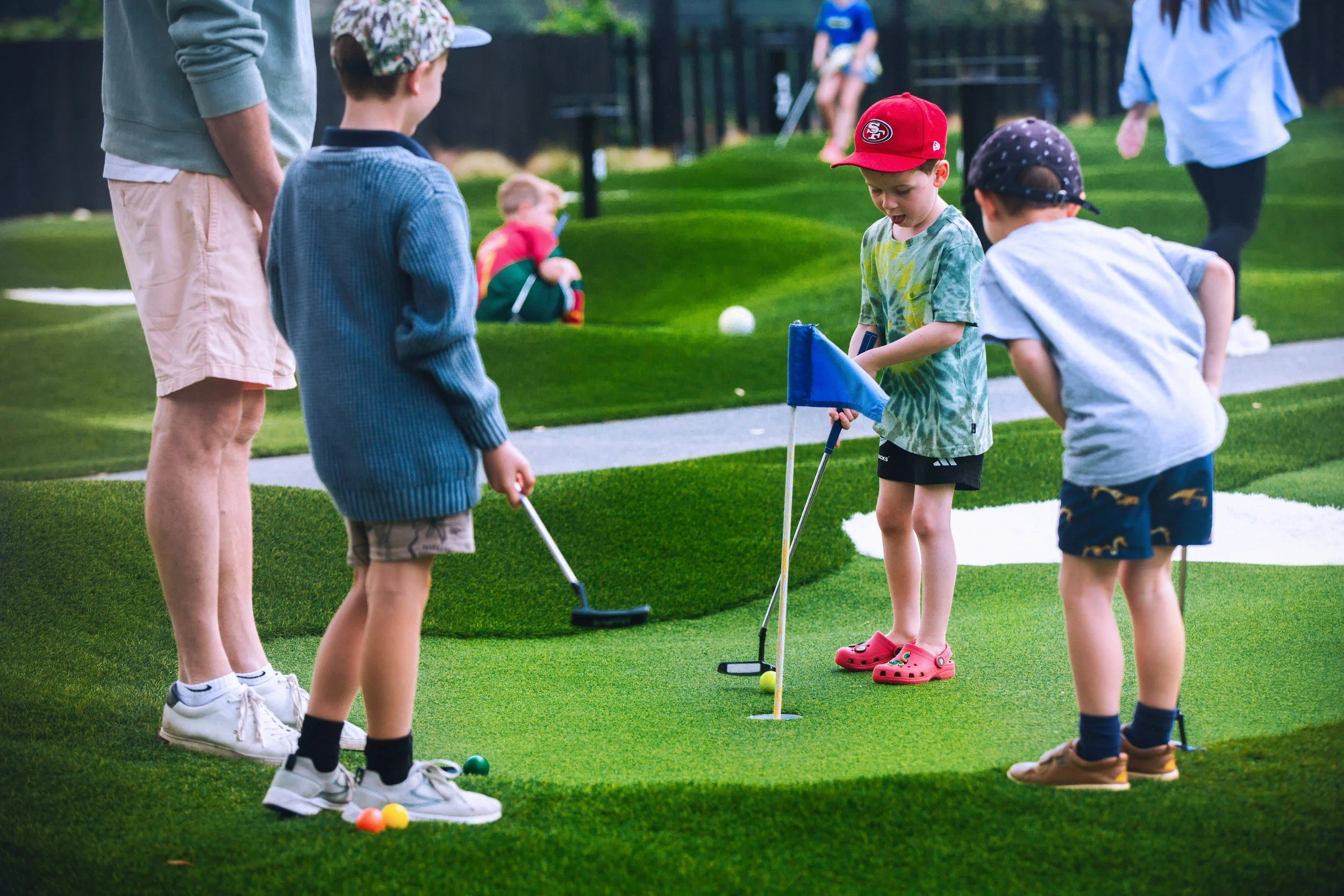 Children playing miniature golf on a putting green, with one boy holding a putter and the others observing, in a park setting.