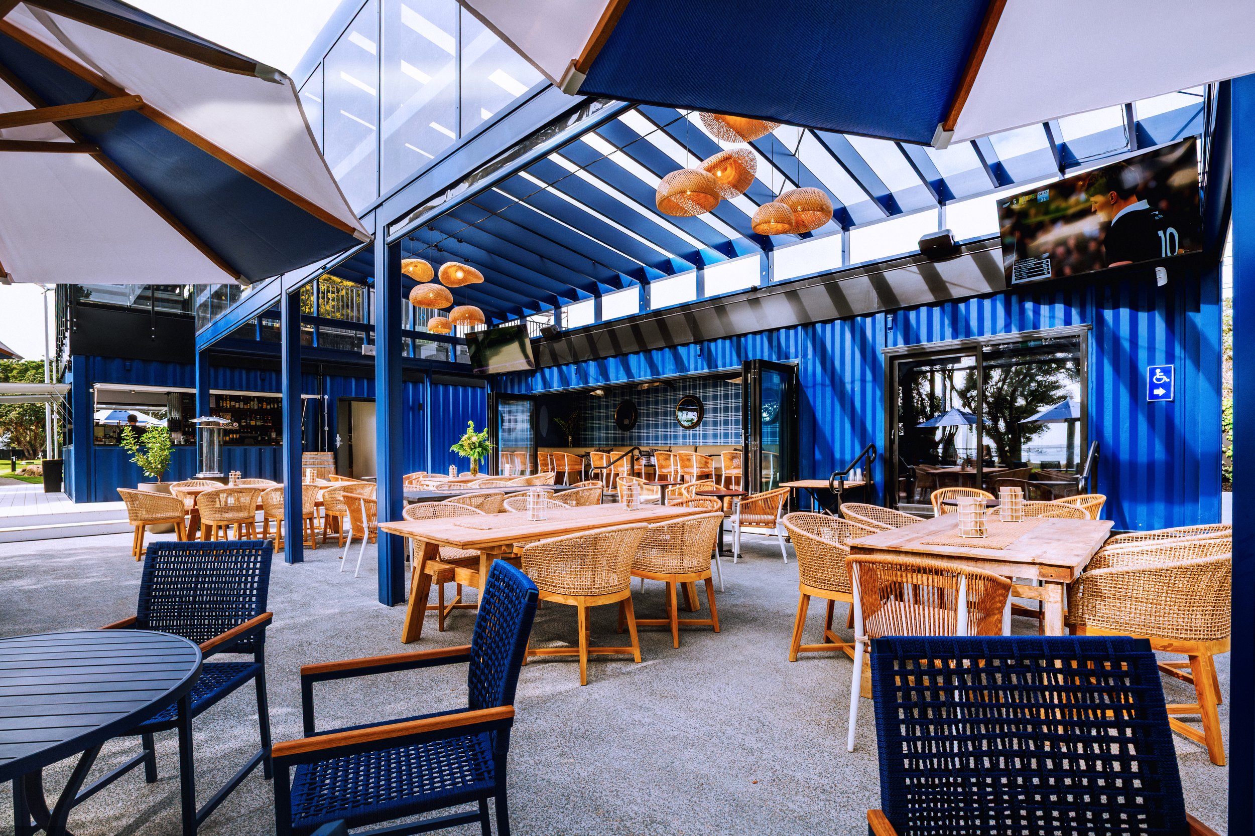 Outdoor patio area of a restaurant or cafe with wooden tables and woven chairs under a blue steel and glass awning, with hanging pendant lights and a large flat-screen TV on the wall.