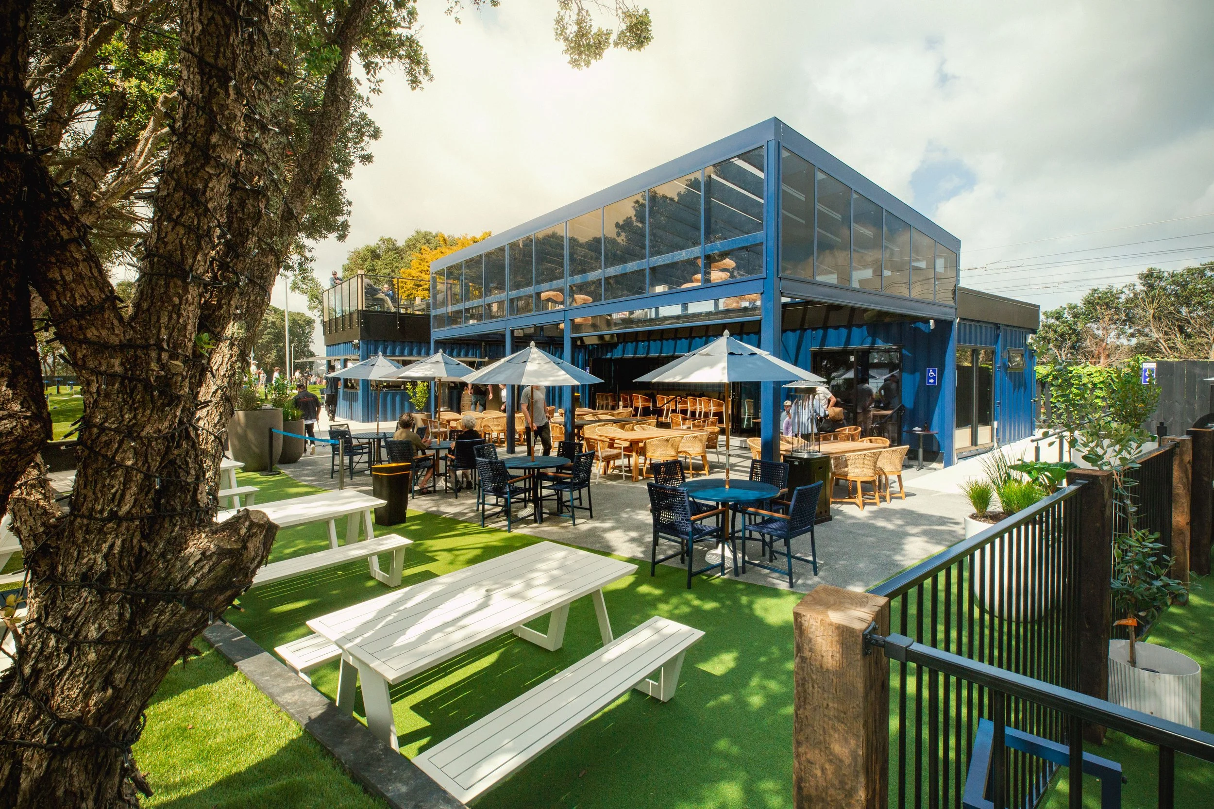 Outdoor patio area with white benches, black and wooden tables, and umbrellas, adjacent to a blue glass-fronted building with two stories, surrounded by trees and greenery on a partly cloudy day.
