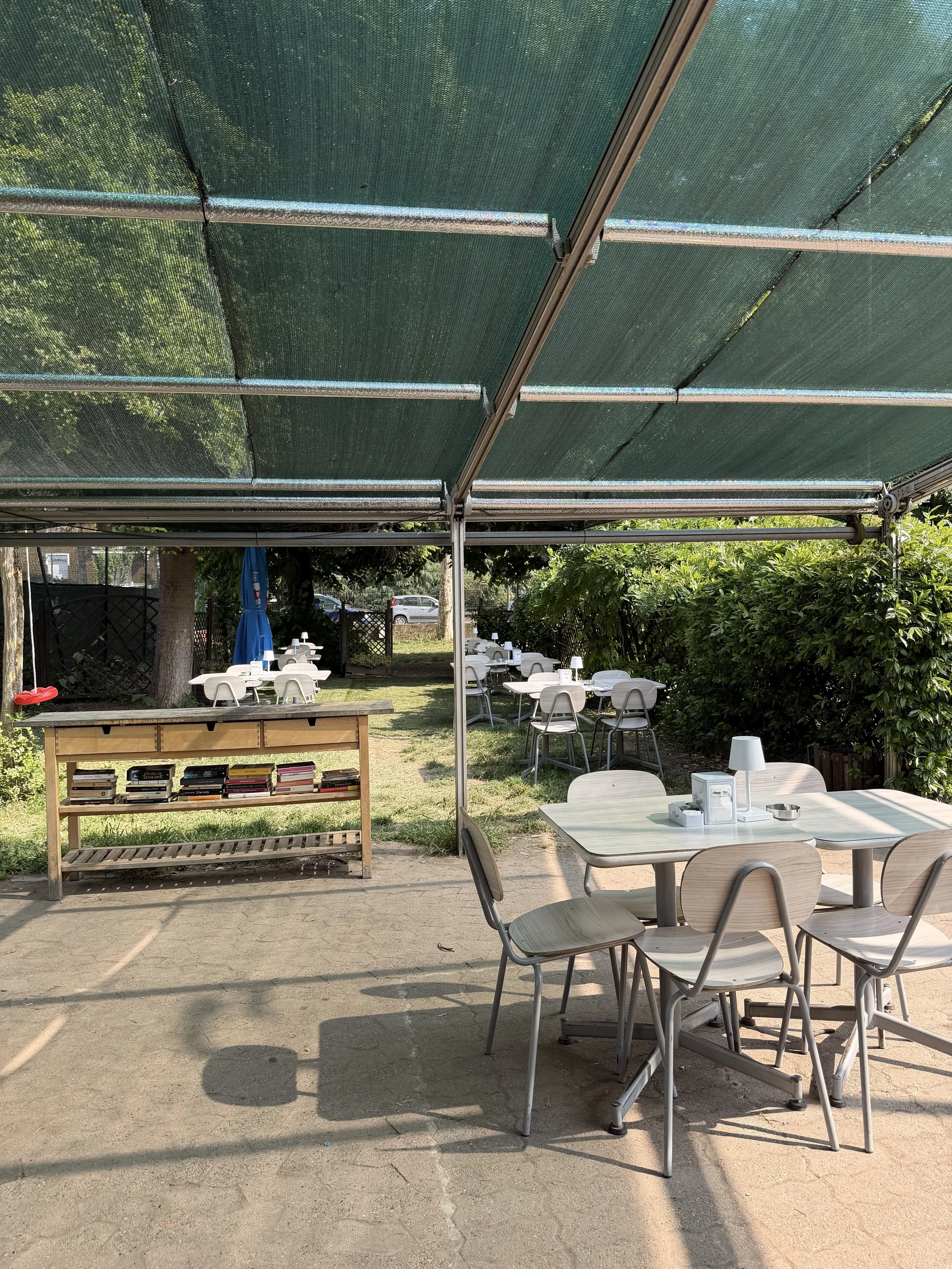 Tavolo e sedie in un ristorante all'aperto sotto un'ombraia, con piante verdi e auto parcheggiate sullo sfondo.