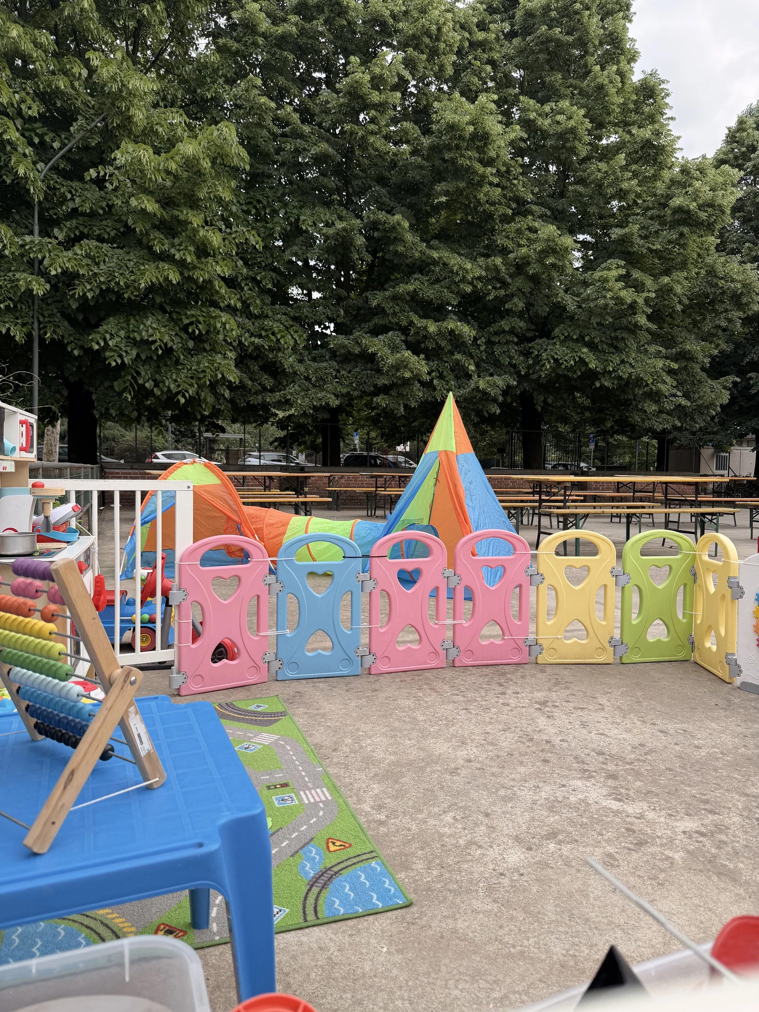 Area giochi all'aperto con giochi colorati, recinto di plastica, tappetini con disegni e alcune strutture di gioco, circondata da alberi e panchine.