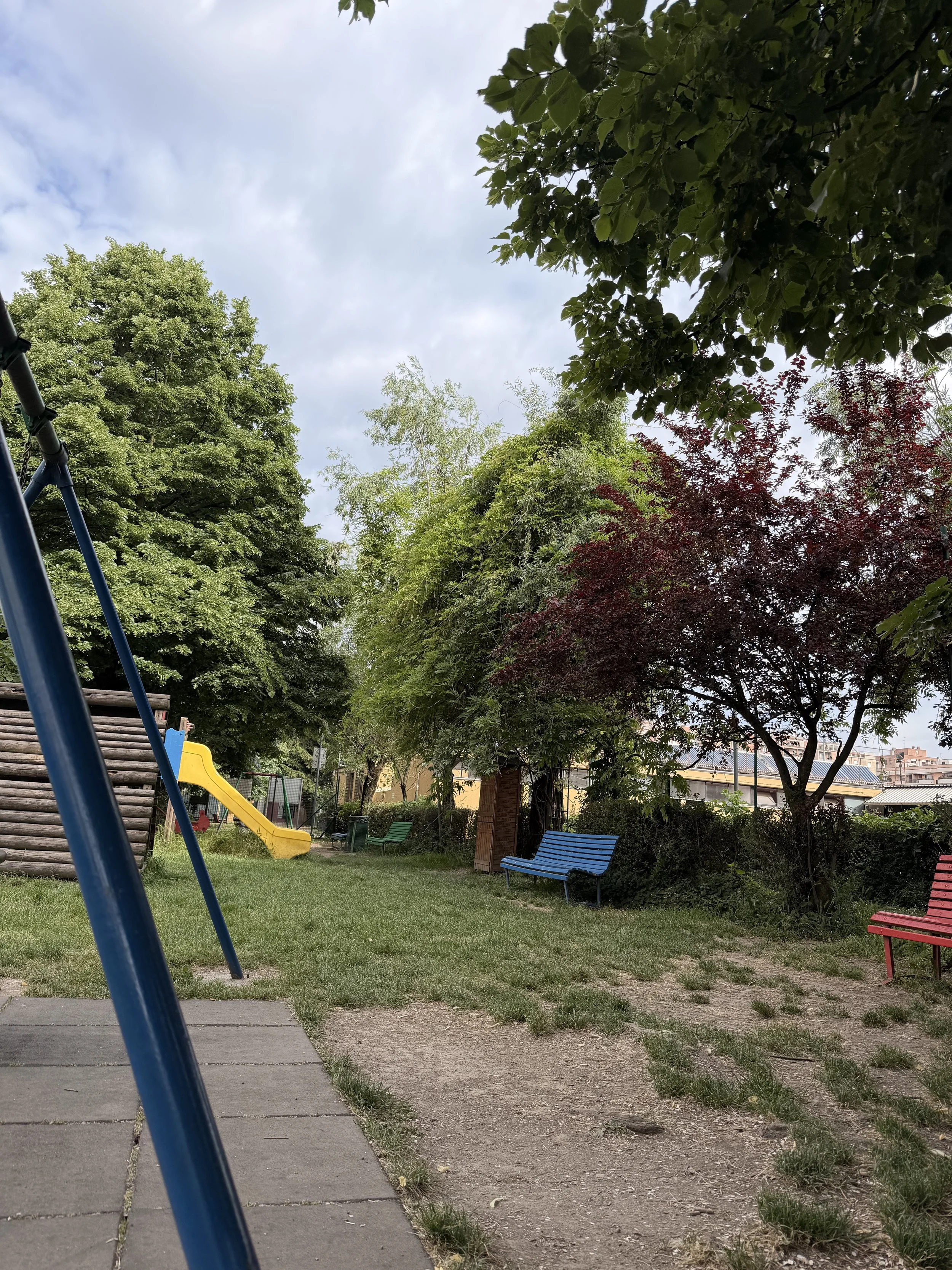 Un parco con altalene, panchine colorate, alberi verdi e un cielo nuvoloso.