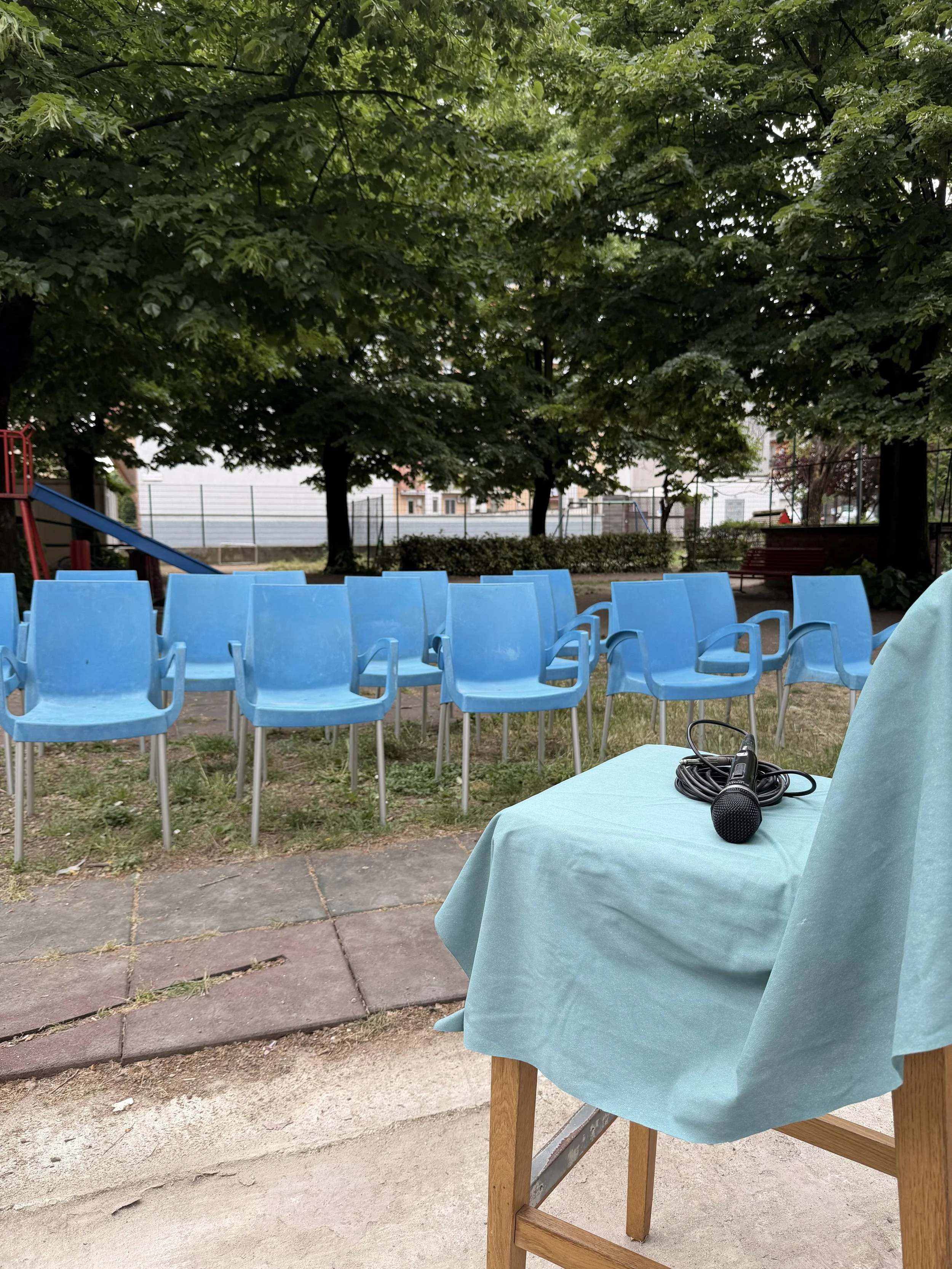 Spettacolo di musica all'aperto con microfono su tavolo coperto da tovaglia azzurra, in un'area verde con sedie blu disposte in cerchio e grandi alberi di fronte a un campo di gioco.
