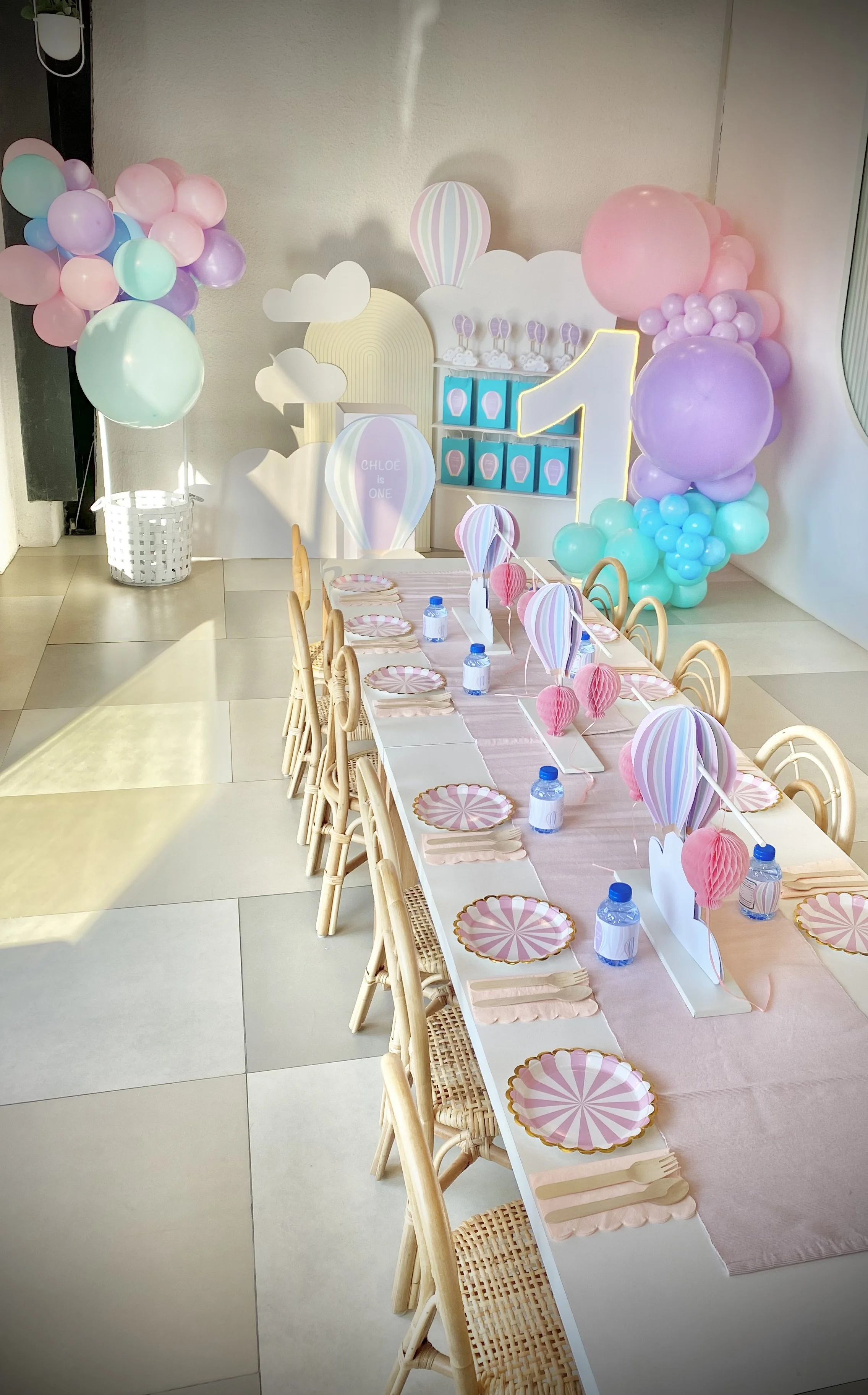 Party setup with pastel-colored balloons, a long table with pink table runner, paper plates and napkins, water bottles, and a backdrop with balloons and clouds for a first birthday celebration.