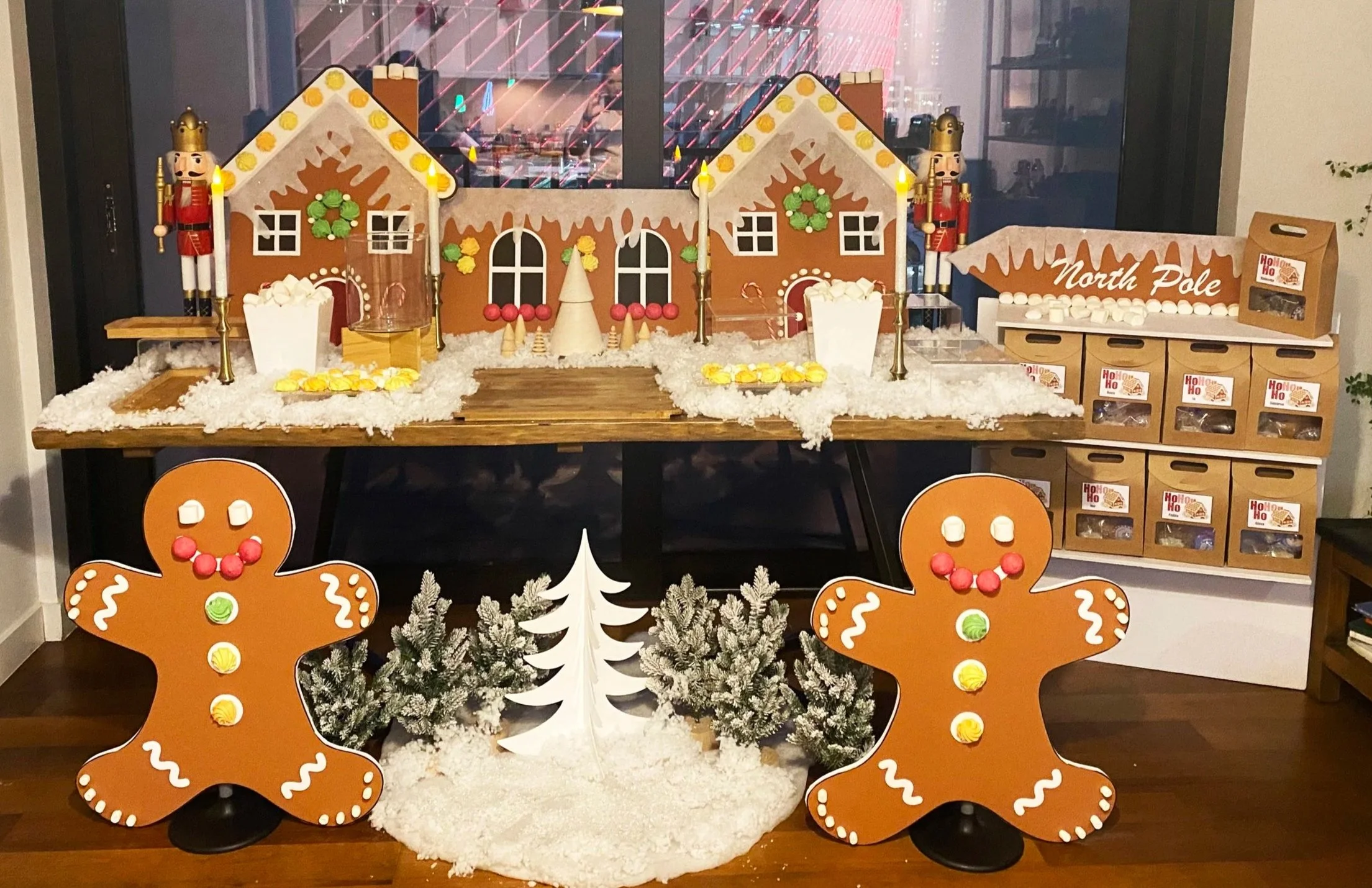 A festive holiday display featuring gingerbread house props, including two gingerbread men, a Christmas tree, and candy-themed decorations, set on a table with artificial snow and Christmas ornaments, with window showing city lights in the background.