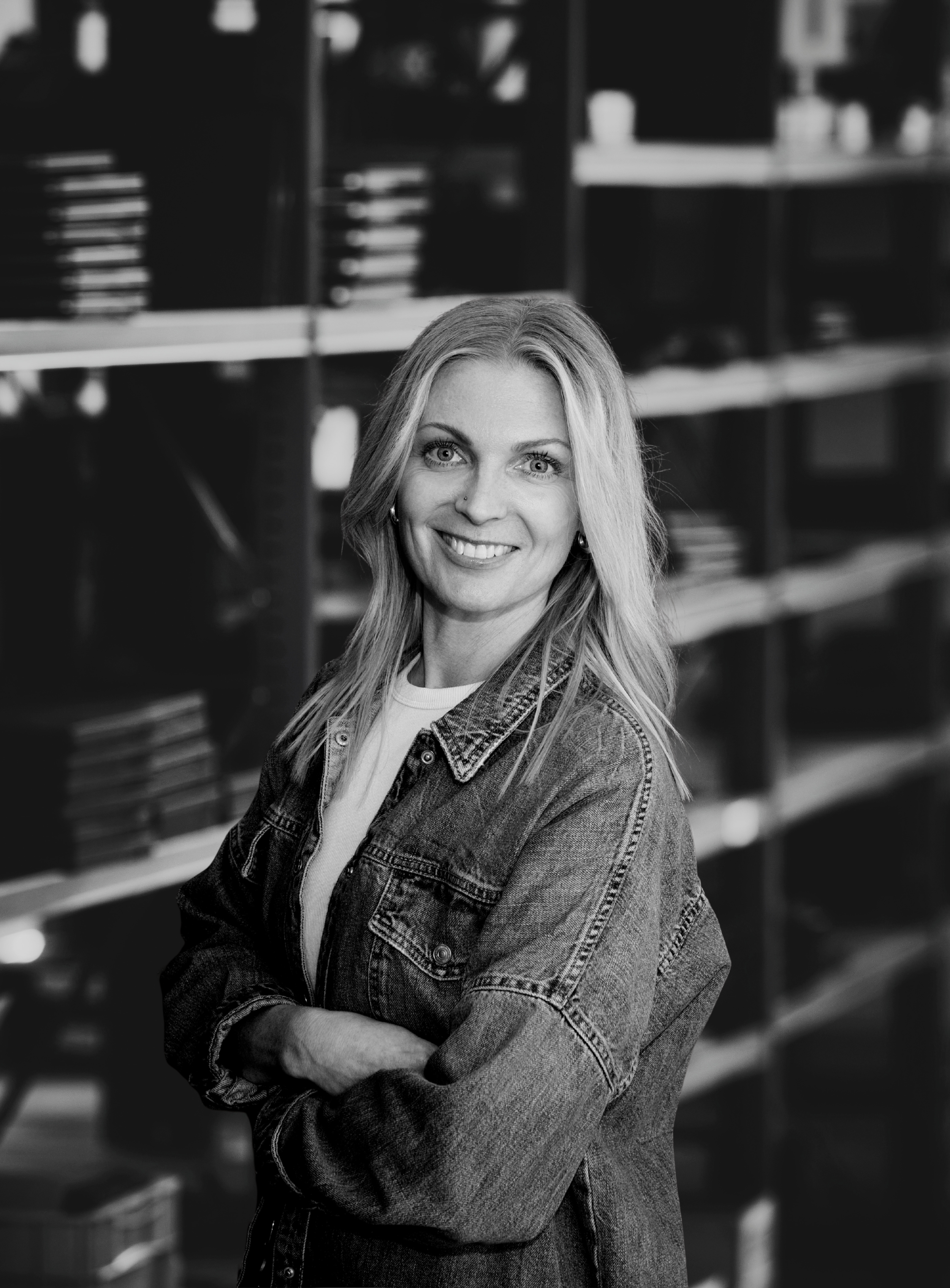 Sort-hvidt fotografi af en ung blonder kvinde med smilende ansigt, iført denimjakke, står med armene krydset foran en bibliotekshylde fyldt med bøger.