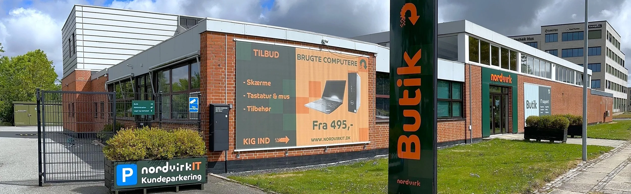 Ude foran en pc butik med grønne og røde skilte, reklame for computere og tilbehør, parkeringsområde med skilt, glasdør indgang, busk og vej på en græsplet.