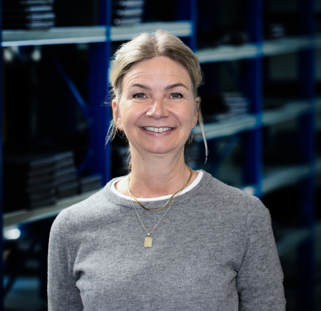 En smilende kvinde med blondt hår og gråt tøj står i en opbevaringshal med blå reoler i baggrunden.