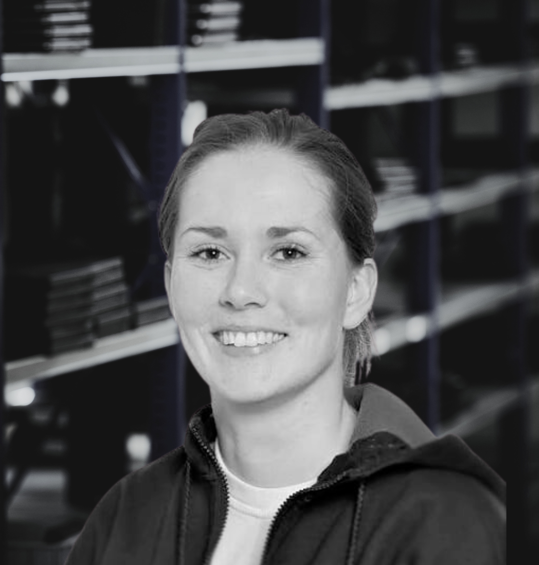 Sort-hvid portræt af en ung kvinde med skulderlangt hår, smilende, klædt i en jakke, foran hylder med filer.
