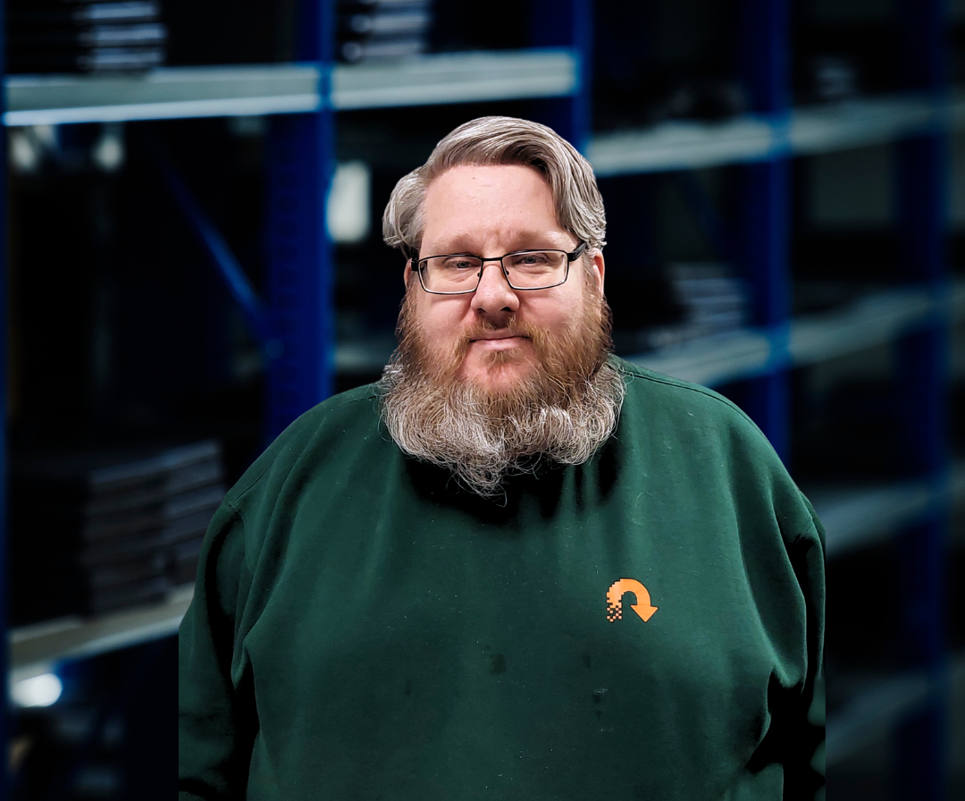 Mand med brunt skæg, briller og grøn sweatshirt stående i et lager.