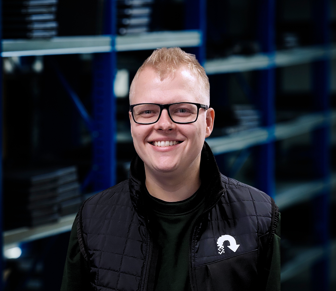 Person med brunt hår, briller, smilende, sort jakke, baggrund med lagerrækker.
