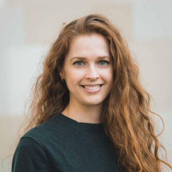 A woman with long, curly red hair, blue eyes, and light skin, smiling and wearing a dark shirt.