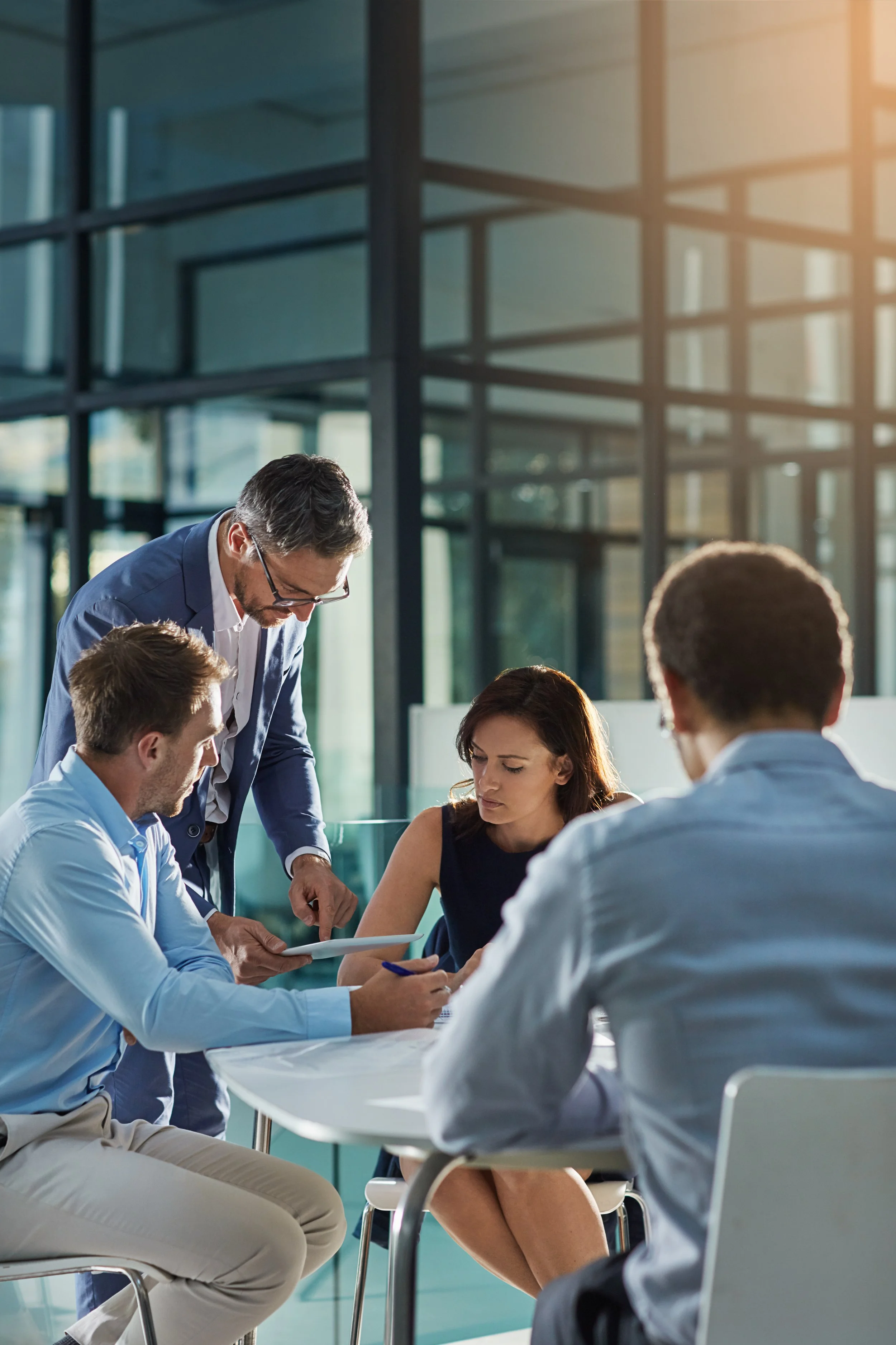 A group of professionals, including a man in a blue suit and glasses, gathered around a table in a modern office building with large glass windows, engaged in a discussion or collaborative work.