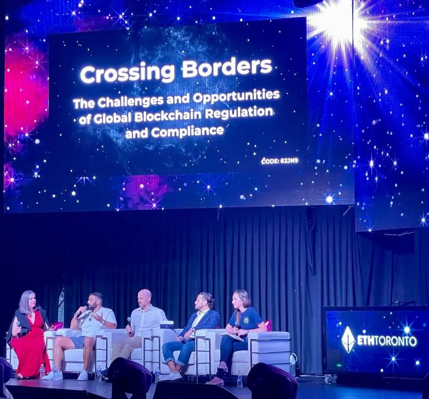 Panel discussion at a conference titled 'Crossing Borders: The Challenges and Opportunities of Global Blockchain Regulation and Compliance' with four people seated on stage, two women and two men, with a large digital screen displaying the title and theme above them.