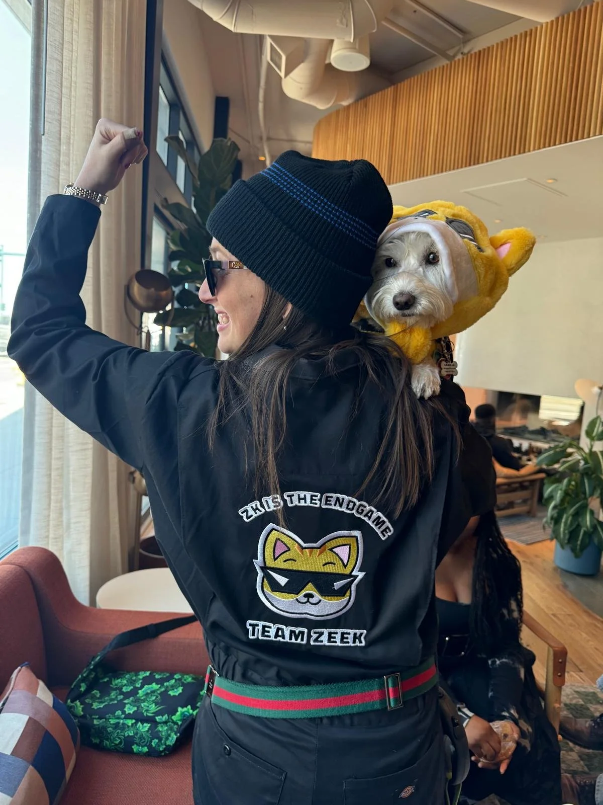 A woman in sunglasses and a beanie with a small dog in a yellow bear costume on her shoulder, taking a selfie in a cafe with light streaming in from a window.