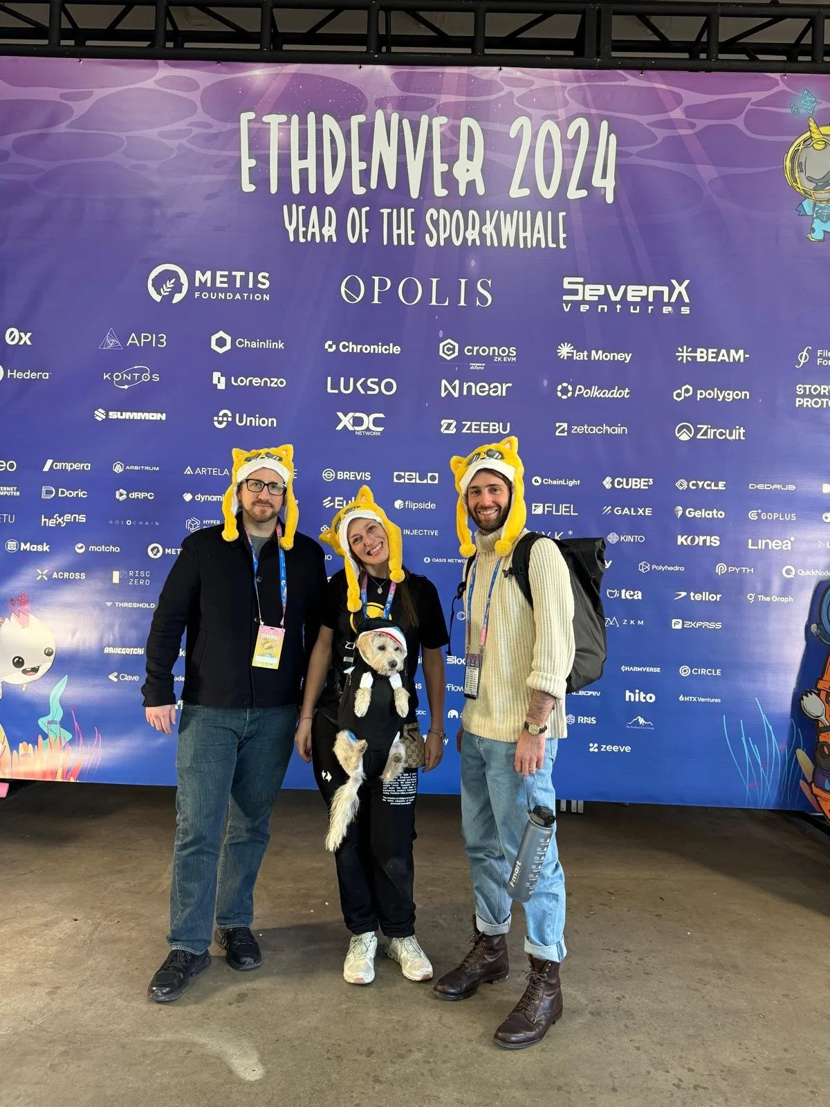 Three people and a dog stand in front of a large purple banner at ETHDenver 2024. They are wearing matching yellow bear-themed hats and smiling, with the two men wearing backpacks and one woman holding the dog.