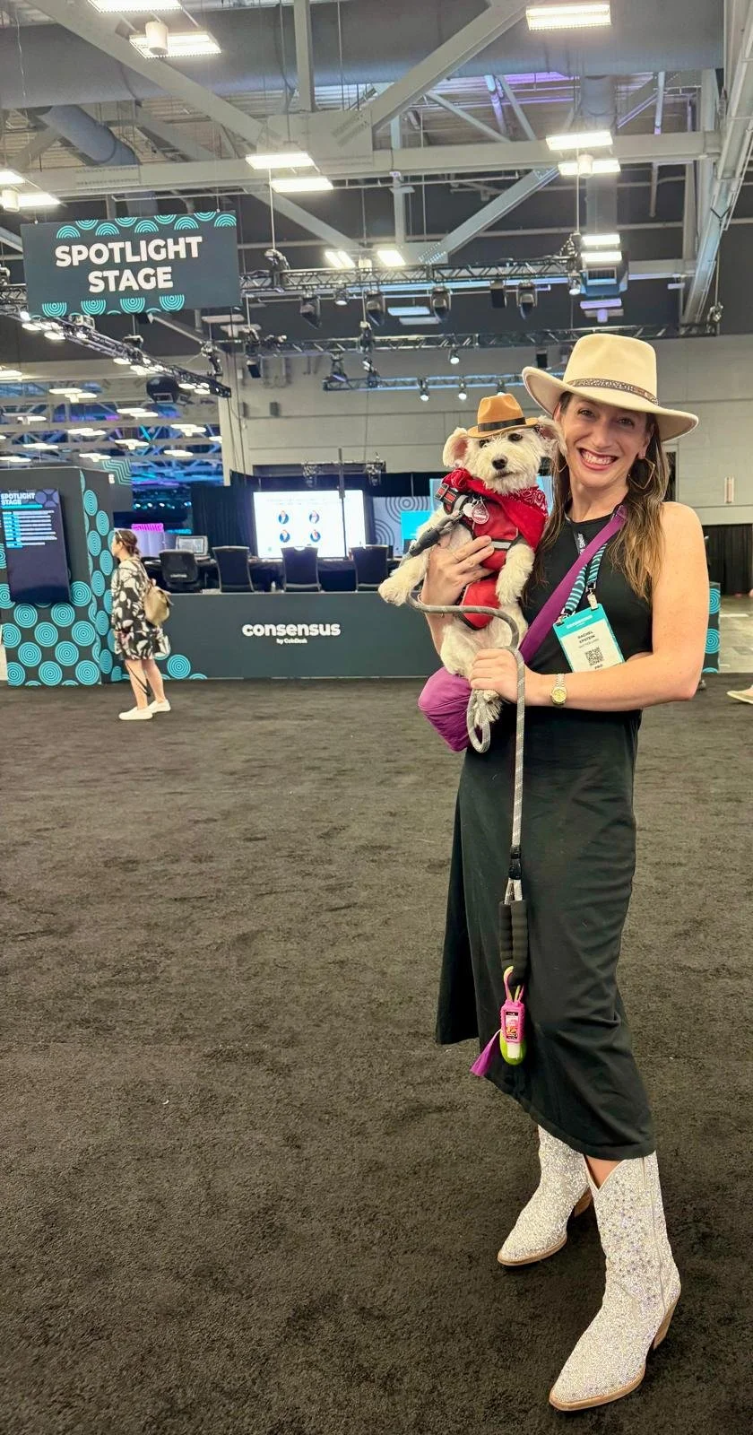 A woman wearing a wide-brimmed hat, black dress, glittery boots, and holding a small dog dressed in a red bandana and harness, standing in an indoor event space with a stage labeled 'Spotlight Stage' and a sign that says 'Consensus' in the background.