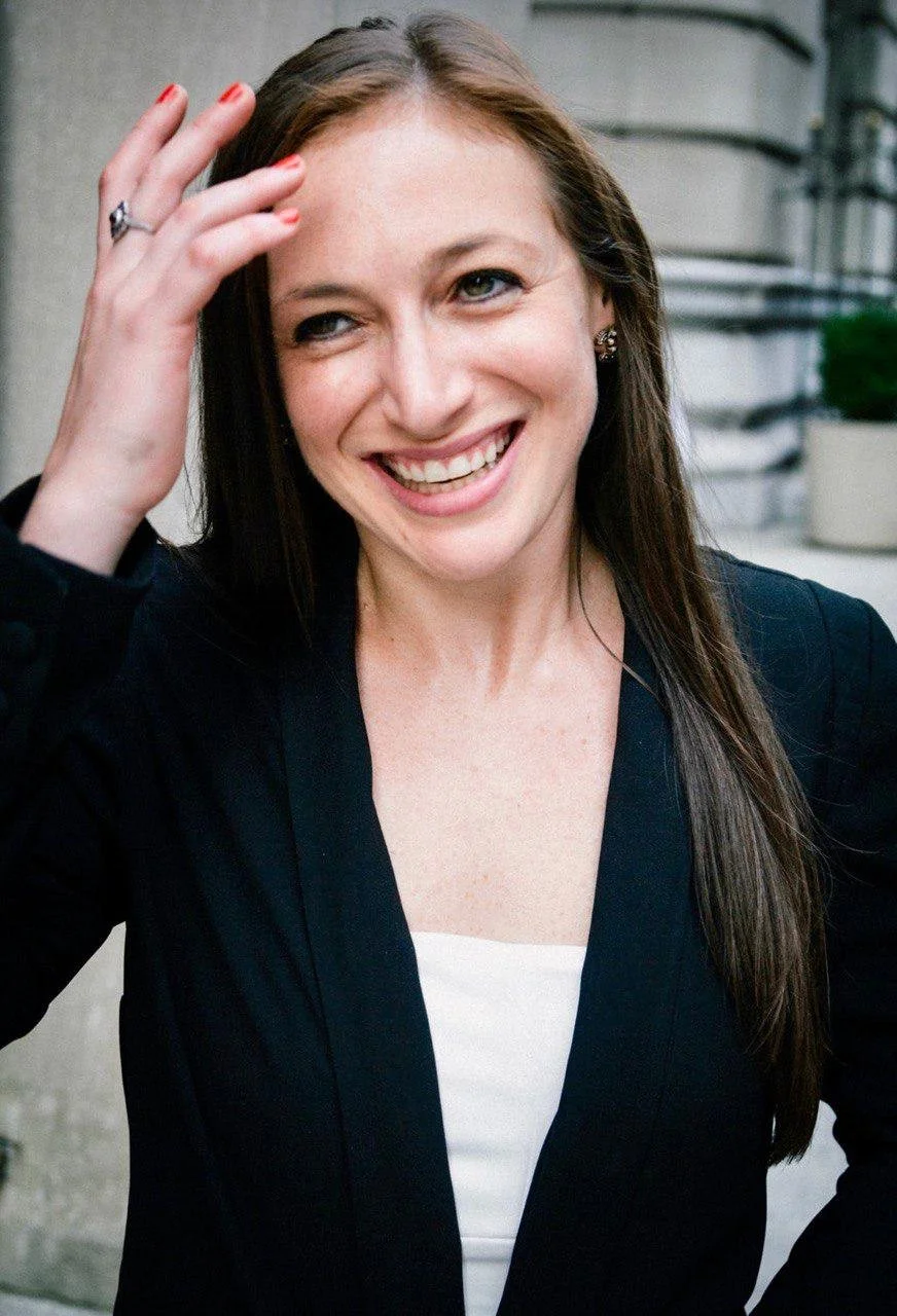 A smiling woman with long brown hair, wearing a black blazer and white top, adjusting her hair outside in front of stone stairs and a planters with green plants.