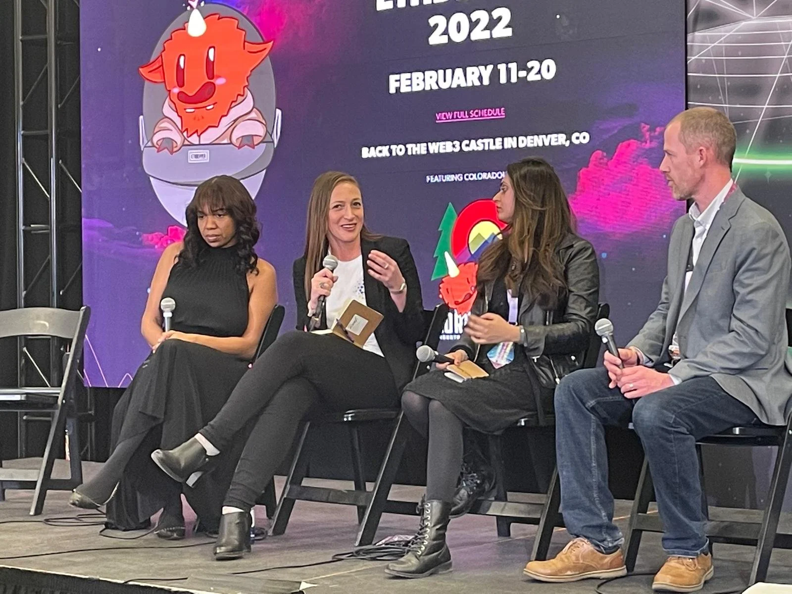 Four people seated on a stage in front of a large screen, participating in a panel discussion at a conference. The screen displays a cartoon lion mascot and event details.