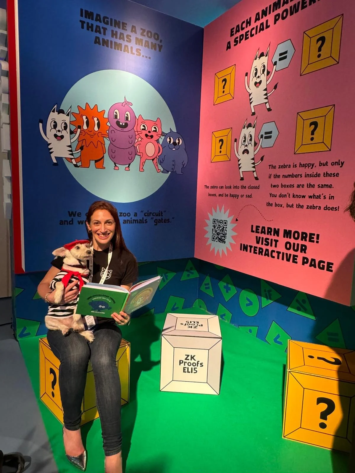A woman smiling and holding a dog dressed in a superhero costume, reading a colorful educational display about animals and number puzzles at a zoo exhibit.